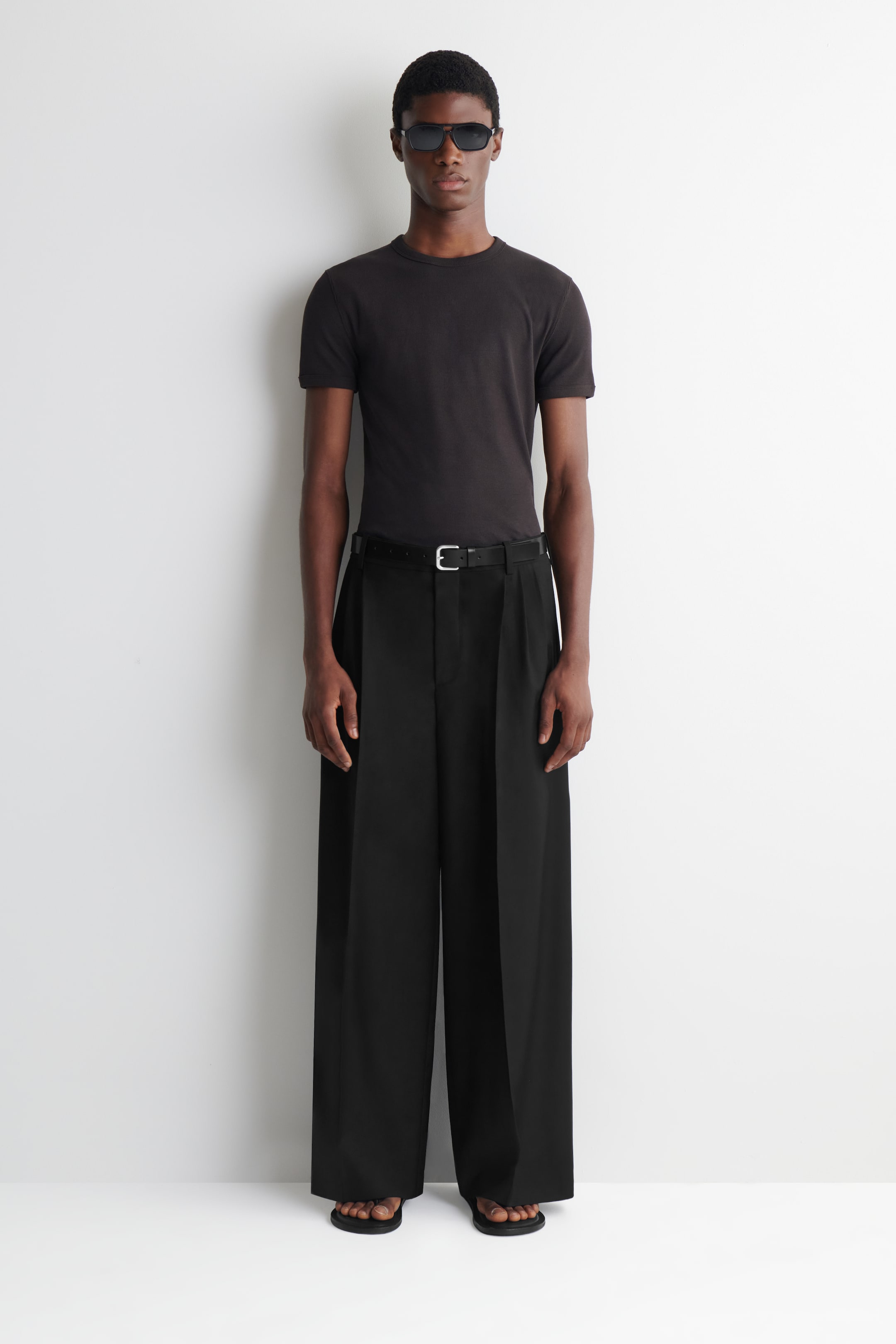 Se større billede: Sorte bukser med brede ben har en høj talje og markante frontlæg, med bæltestropper der sikrer et sort bælte med et sølvspænde. Deres fuldlængde snit skaber en flydende, bred silhuet.