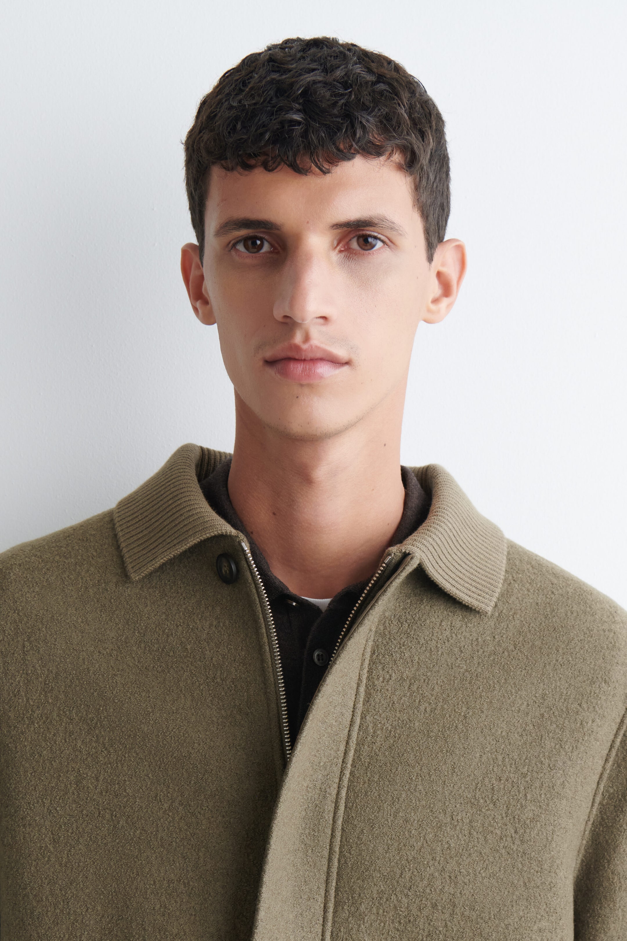 The model is wearing a ribbed-collar boiled-wool chore jacket in an olive-green tone, featuring a textured wool body, a ribbed knit collar, and a partially open metal zipper. The jacket is layered over a dark brown buttoned polo shirt. The model is wearing a ribbed-collar boiled-wool chore jacket in an olive-green tone, featuring a textured wool body, a ribbed knit collar, and a partially open metal zipper. The jacket is layered over a dark brown buttoned polo shirt.