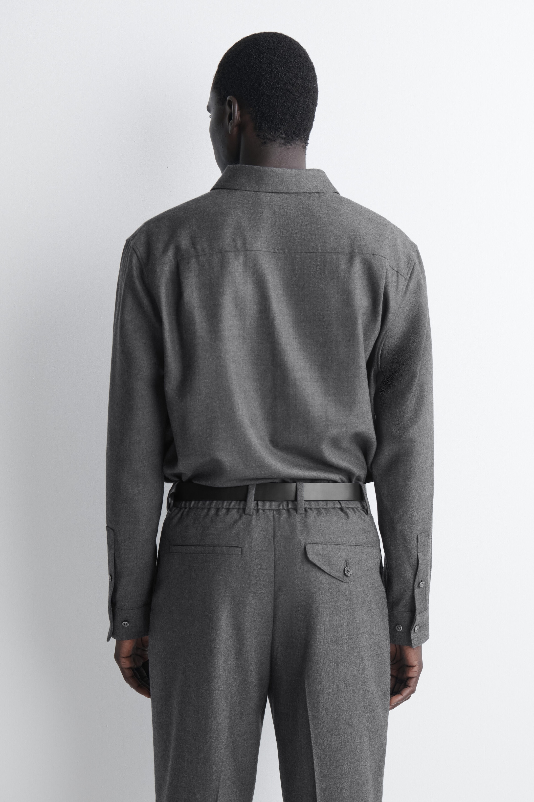 Das locker geschnittene Hemd aus Wollflanell ist in einer Rückansicht zu sehen und präsentiert eine klare horizontale Passe sowie lange Ärmel mit geknöpften Manschetten. Das Hemd wird in die dazu passende graue Hose mit schwarzem Gürtel gesteckt getragen, wodurch seine weiche Textur und der entspannte Fall betont werden. Das locker geschnittene Hemd aus Wollflanell ist in einer Rückansicht zu sehen und präsentiert eine klare horizontale Passe sowie lange Ärmel mit geknöpften Manschetten. Das Hemd wird in die dazu passende graue Hose mit schwarzem Gürtel gesteckt getragen, wodurch seine weiche Textur und der entspannte Fall betont werden.