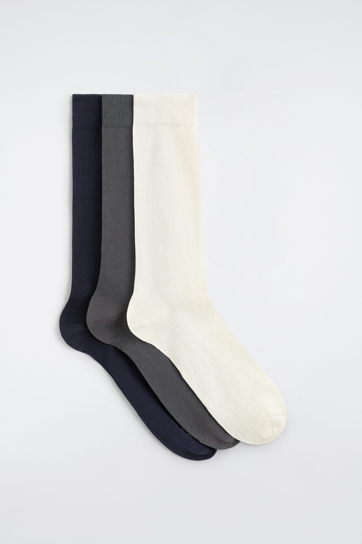 Un lot de trois paires de chaussettes en coton mercerisé est disposé à plat dans une composition empilée et décalée, mettant en valeur des coloris bleu marine, gris foncé et blanc cassé de profil. Les chaussettes présentent une finition lisse en maille fine, avec des bords côtelés en haut.