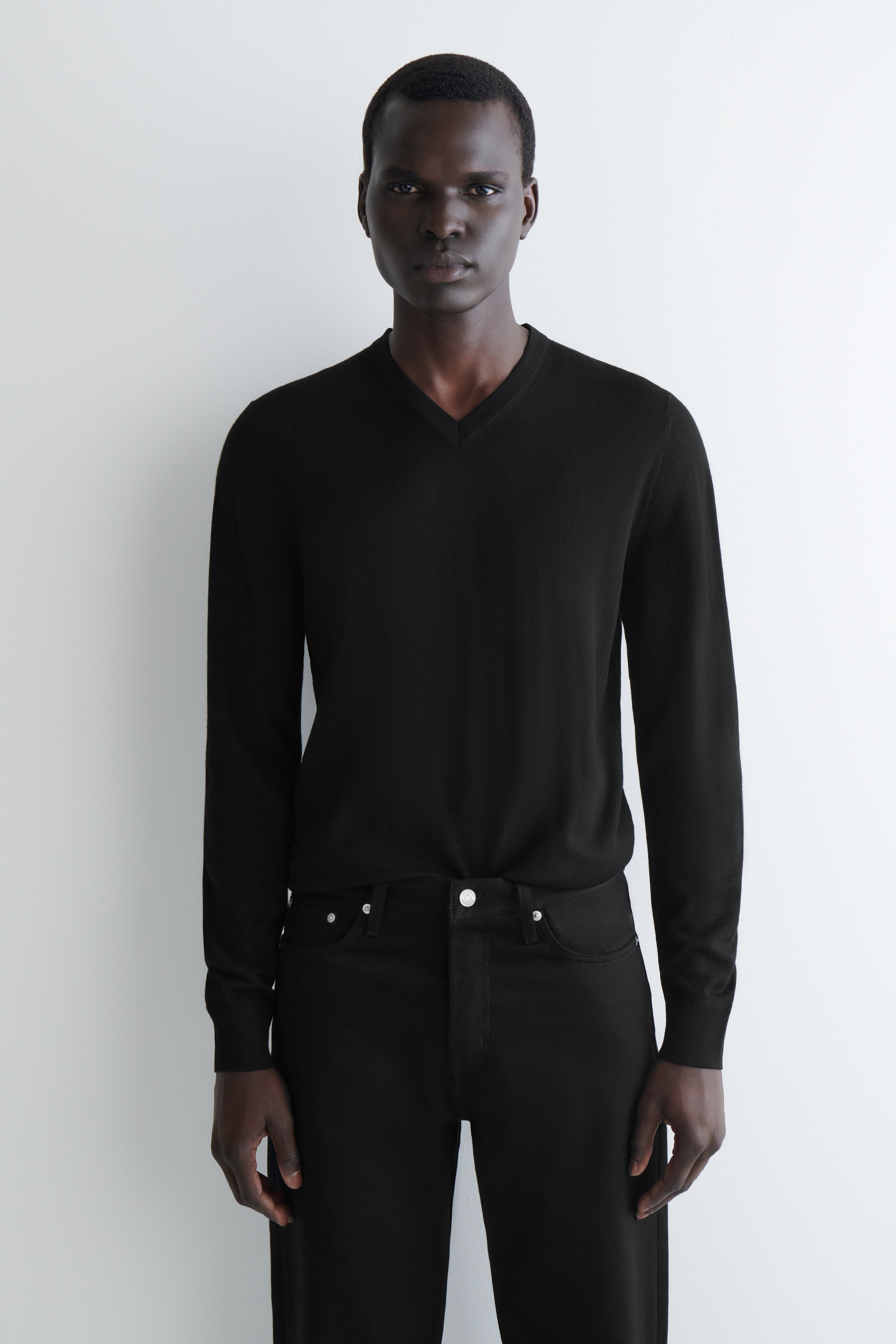 Visualizza immagine più grande: Un maglione nero con scollo a V è presentato frontalmente, caratterizzato da maniche lunghe con polsini a coste e una texture liscia e a maglia fine. Il maglione è indossato infilato in pantaloni neri, mettendo in mostra la sua vestibilità e stile.
