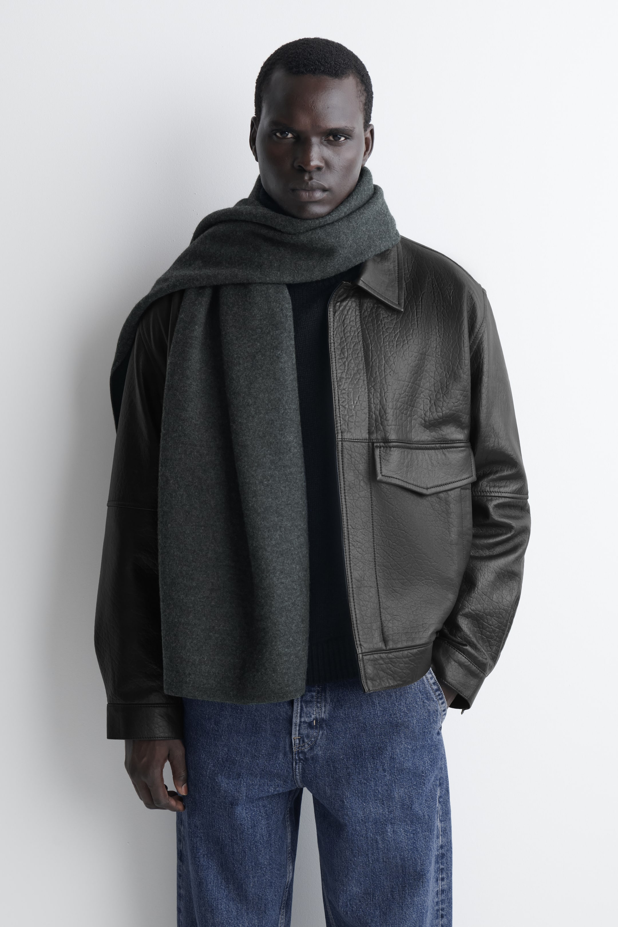 Ver imagen más grande: Una bufanda de lana hervida gris oscuro se enrolla alrededor del cuello de una modelo de frente, con un extremo largo cayendo por la parte delantera de una chaqueta de cuero oscuro. La bufanda muestra una textura suave y densa, con bordes limpios y minimalistas.