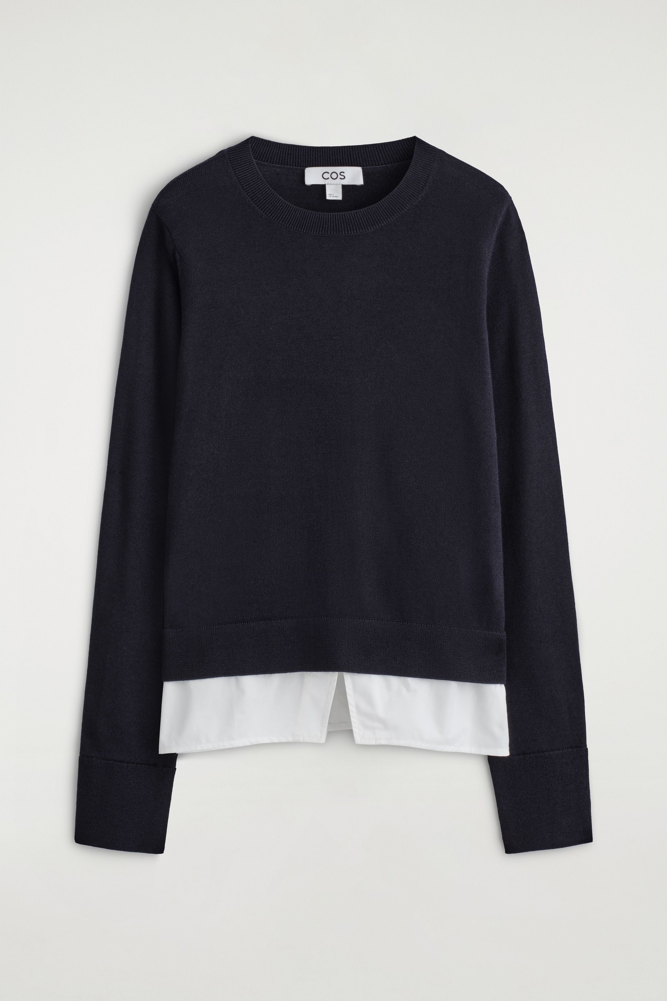 Visualizza immagine più grande: Un maglione blu navy a maniche lunghe viene presentato dalla PARTE ANTERIORE, caratterizzato da un collo a girocollo a coste e polsini a coste. Un pannello di camicia bianca si estende sotto l'orlo, creando un effetto a strati con una spaccatura centrale visibile.