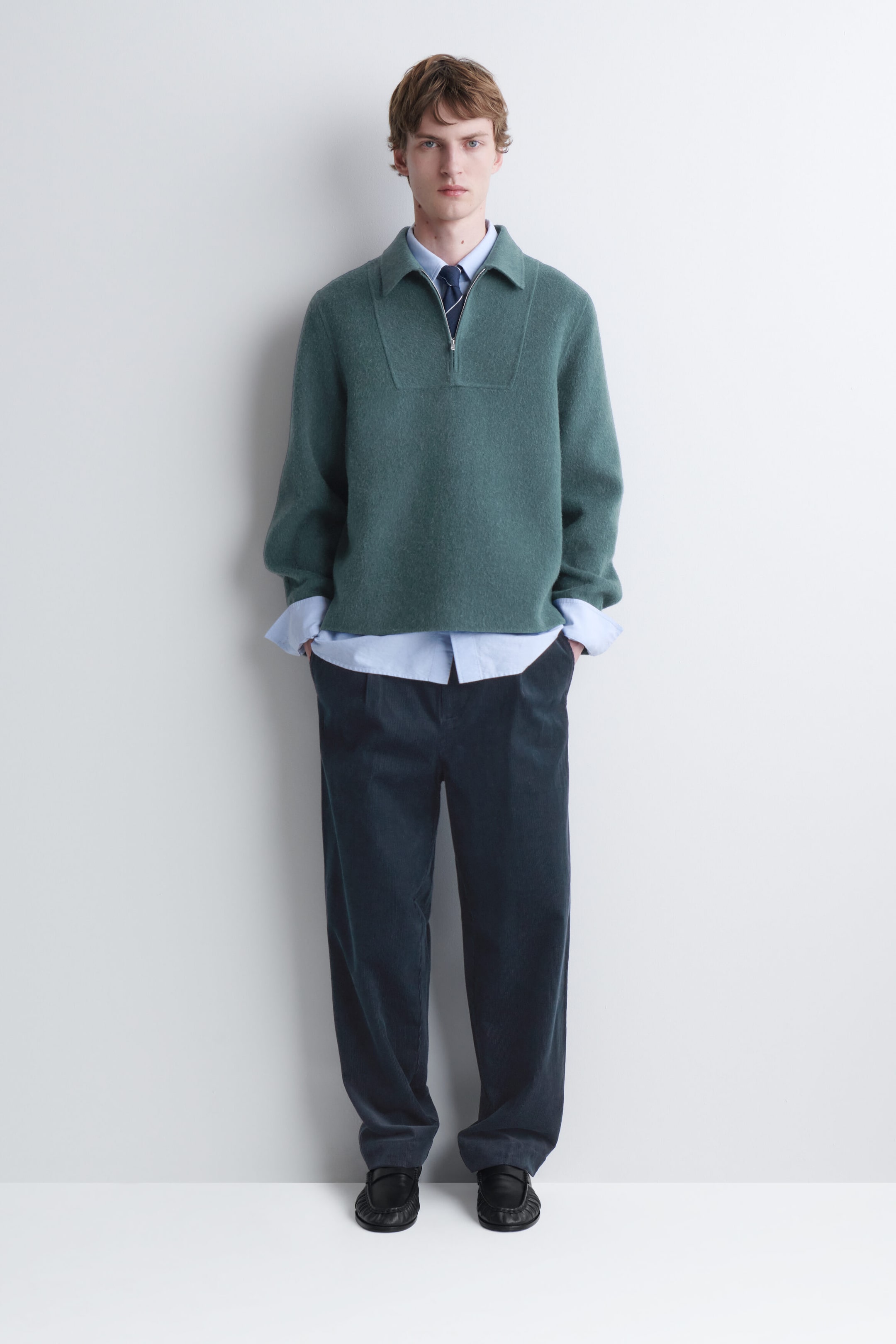A green knit half-zip sweater with a collar is being worn by the model. The sweater is layered over a light blue collared shirt and a dark tie, and is paired with dark blue corduroy pants. A green knit half-zip sweater with a collar is being worn by the model. The sweater is layered over a light blue collared shirt and a dark tie, and is paired with dark blue corduroy pants.