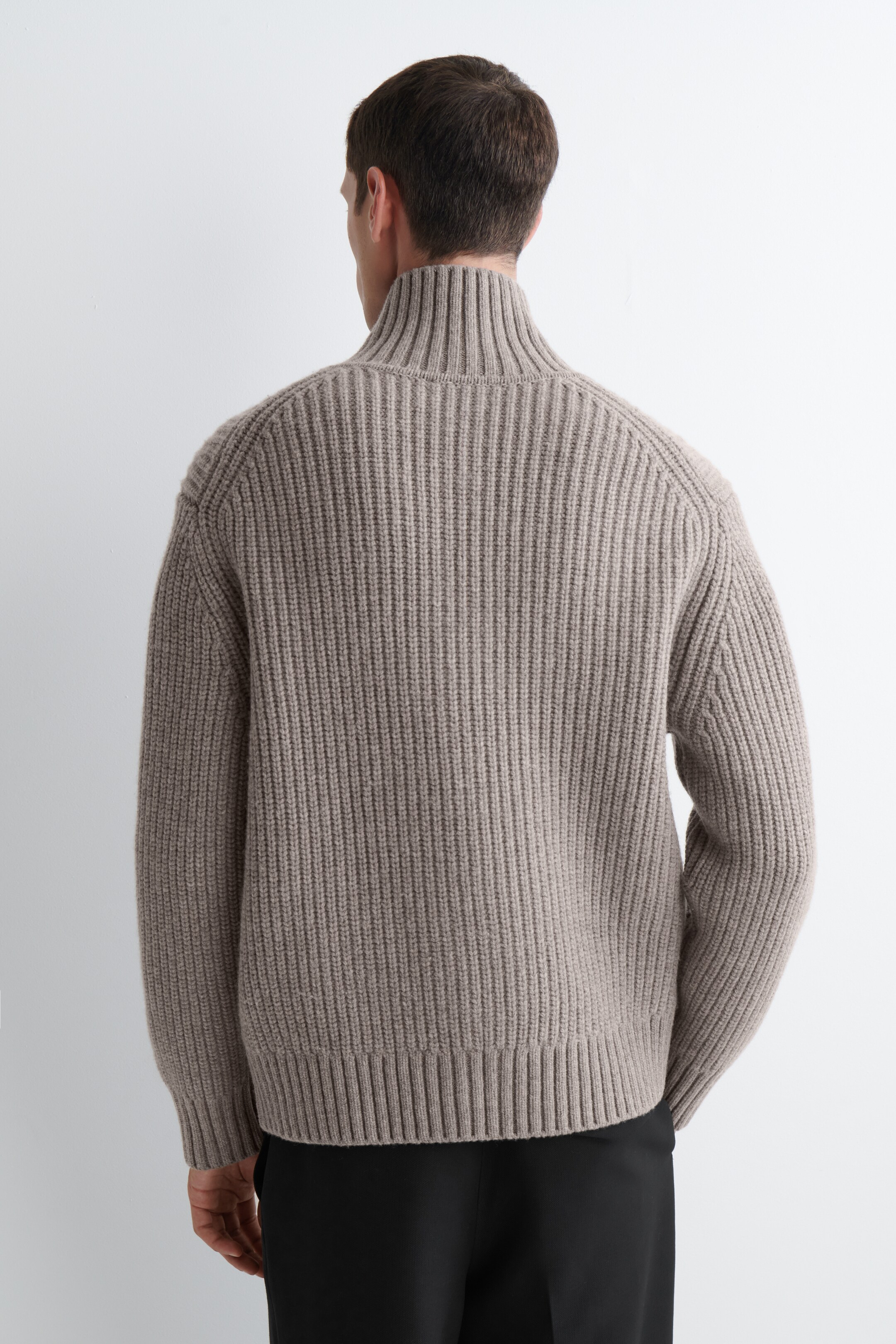 Se større billede: En beige sweater præsenteres bagfra med en tyk ribstrikket struktur overalt. Den er designet med en høj ribbet krave, lange ærmer og en ribbet kant.