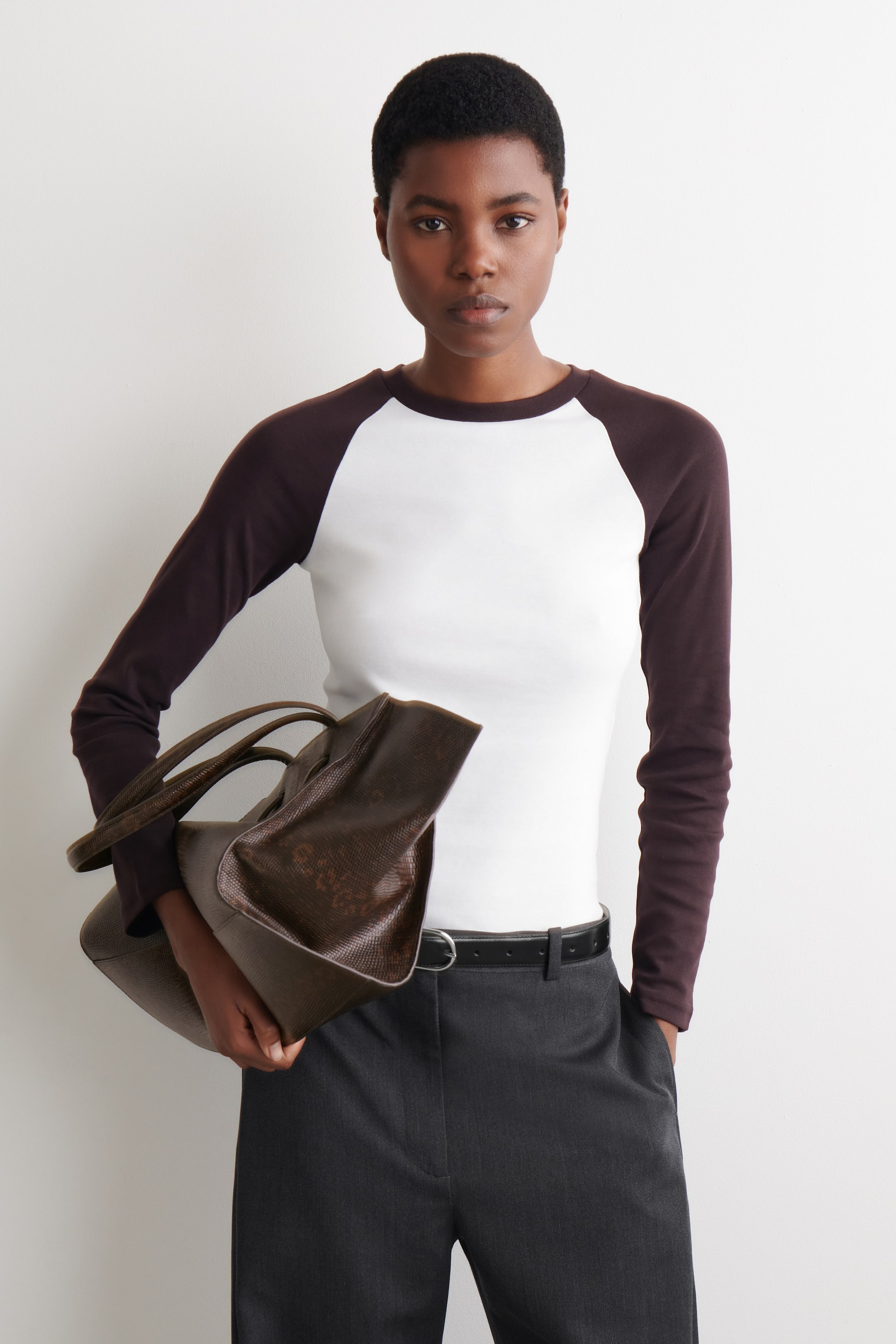 Ingrandisci l'immagine: La modella indossa una T-shirt con corpo bianco e maniche raglan lunghe di colore marrone scuro. Indossa anche pantaloni grigio scuro con una cintura nera e porta una borsa a mano marrone scuro.