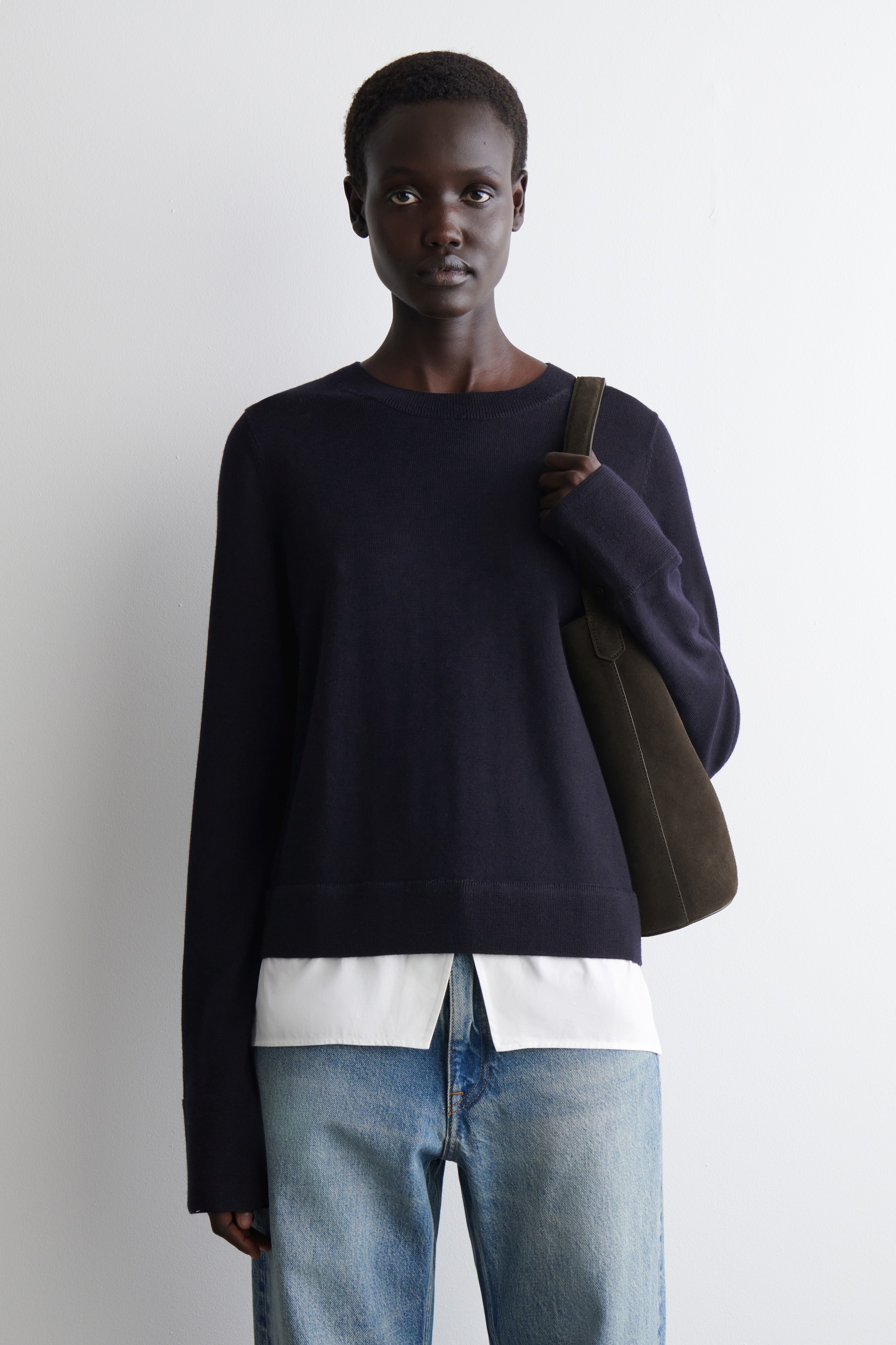 Visualizza immagine più grande: Un maglione girocollo blu navy è indossato, con maniche lunghe, polsini a coste e orlo a coste. È stratificato sopra una camicia bianca.