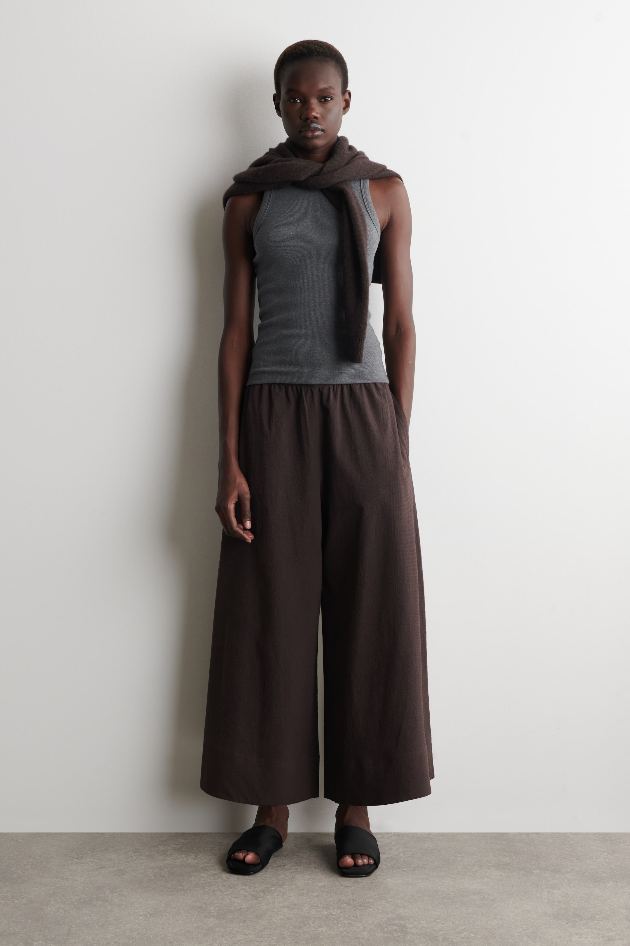 הצגת תמונה גדולה יותר: גופיית טנק אפורה עם צלעות, בעלת מחשוף עגול, נלבשת עם מכנסיים רחבים בצבע חום כהה, עם בד מרקם וגומי במותן, שמסתיימים מעל הקרסוליים. צעיף רך בצבע חום כהה מונח על הכתפיים והחזה, ונעלי סלייד שטוחות בצבע שחור עם רצועה רחבה אחת ננעלות על הרגליים.