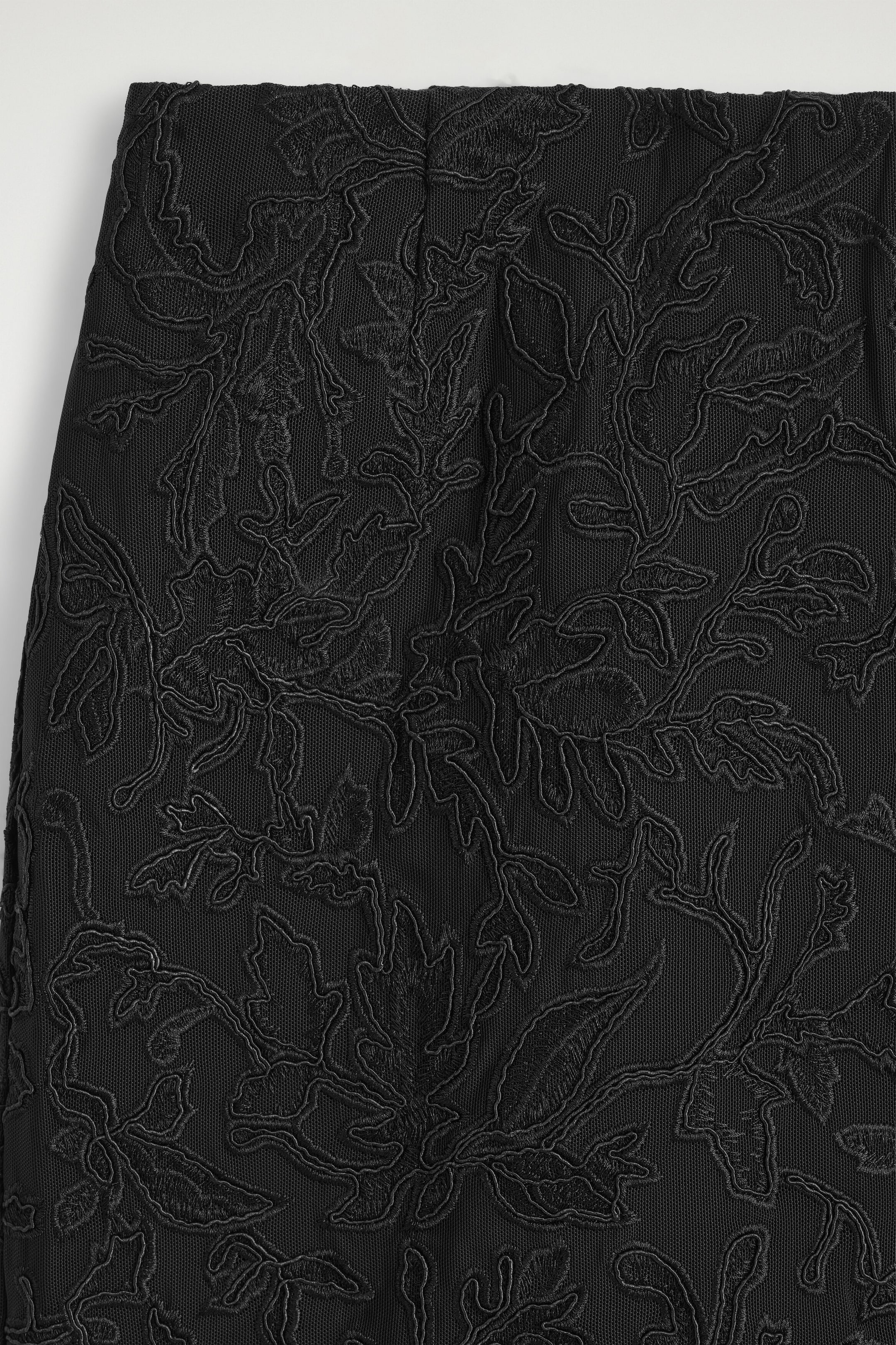 A close-up view shows the textured fabric of a black midi skirt, featuring intricate floral and leaf embroidery layered over a fine mesh backdrop. A close-up view shows the textured fabric of a black midi skirt, featuring intricate floral and leaf embroidery layered over a fine mesh backdrop.