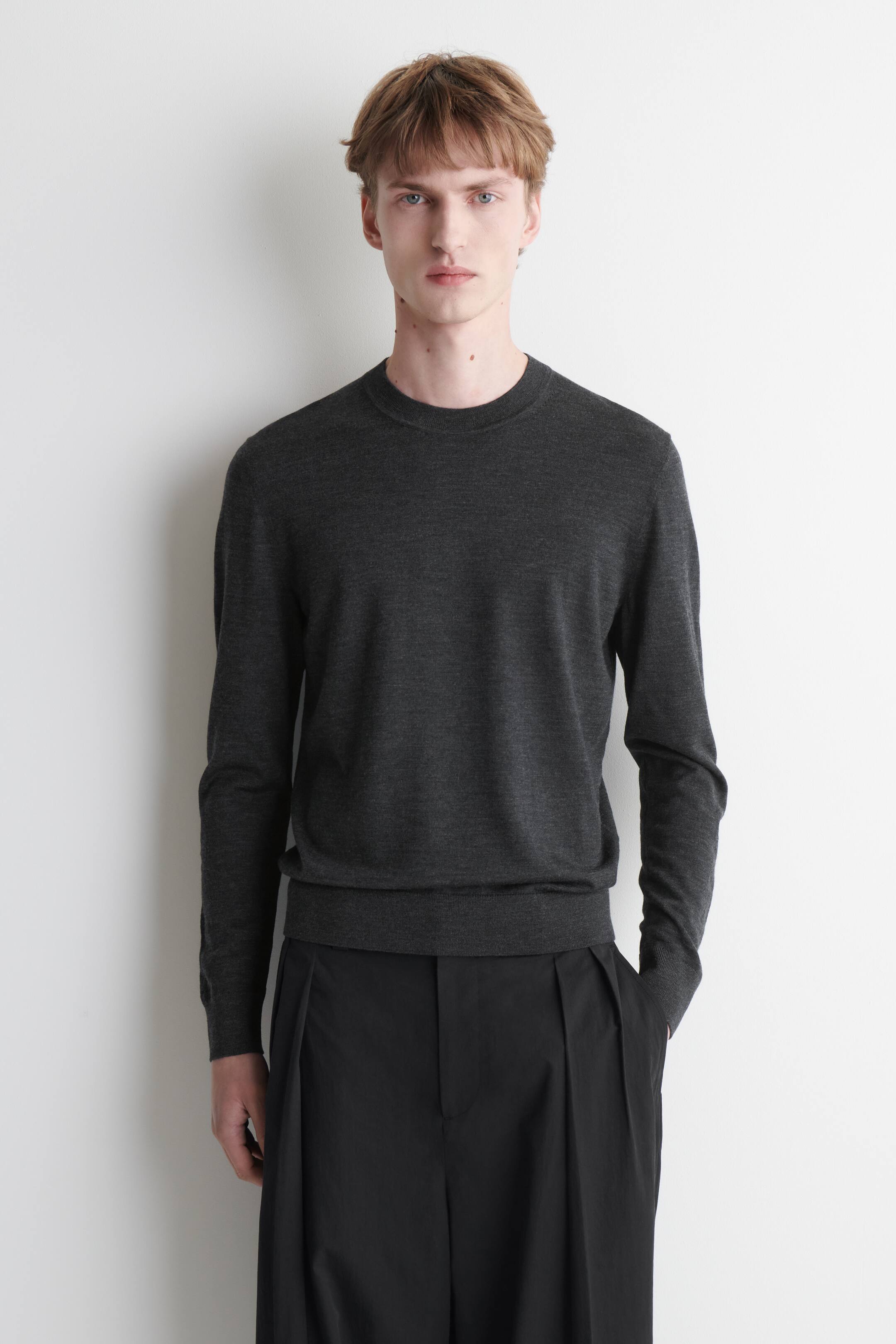 Grösseres Bild ansehen: Ein dunkelgrauer Merino-Wollpullover mit geripptem Rundhalsausschnitt, gerippten Ärmelabschlüssen und geripptem Saum wird vom Model getragen. Der Pullover hat lange Ärmel und ist mit einer schwarzen Hose kombiniert.