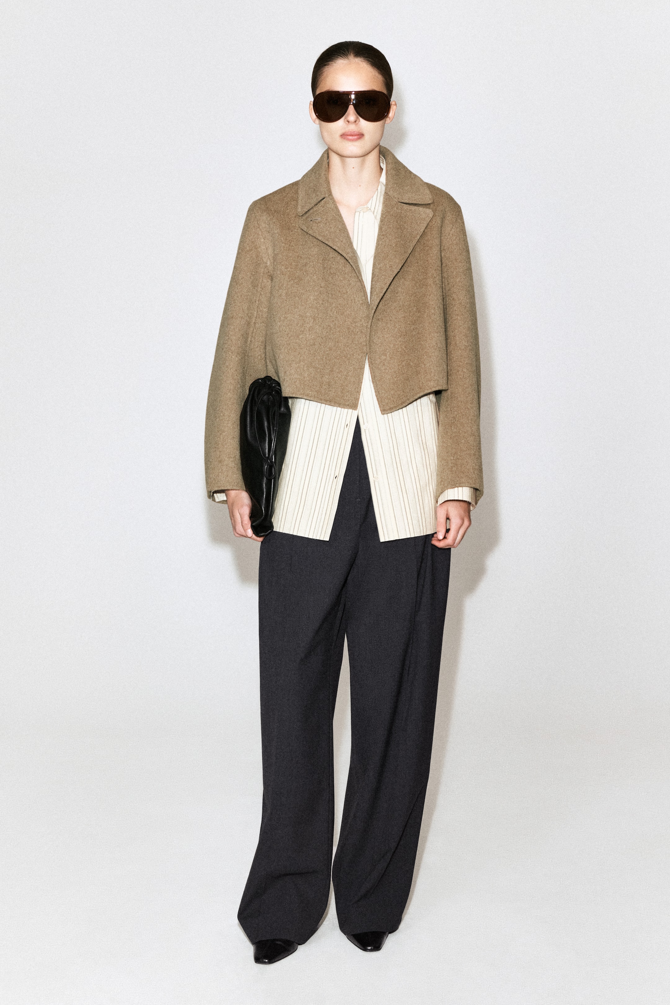 Agrandir l'image: Une veste courte marron clair à larges revers est superposée à une chemise crème à rayures verticales, associée à un pantalon large gris foncé et à des chaussures noires à bout pointu. Une pochette noire froncée est tenue à la main, et de grandes lunettes de soleil noires sont portées.
