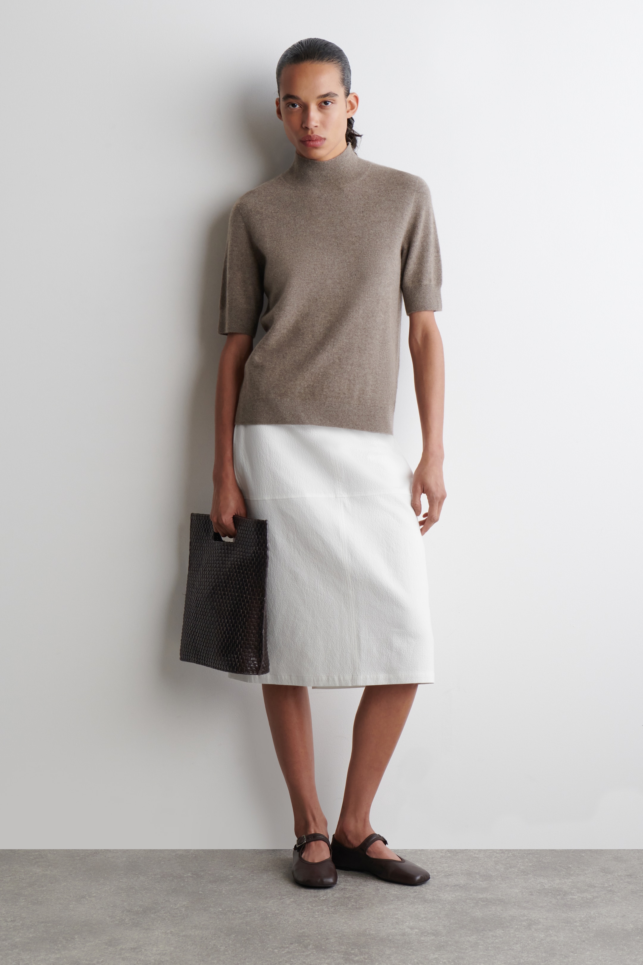 Agrandir l'image: Un ensemble complet est présenté, avec un haut taupe à manches courtes et col montant, de coupe décontractée et à ourlet droit. Une jupe midi blanche texturée avec une couture horizontale est portée, offrant une silhouette droite qui tombe sous les genoux. Un sac à main tressé brun foncé est tenu, et des ballerines Mary Jane brun foncé avec une bride à boucle sont portées.