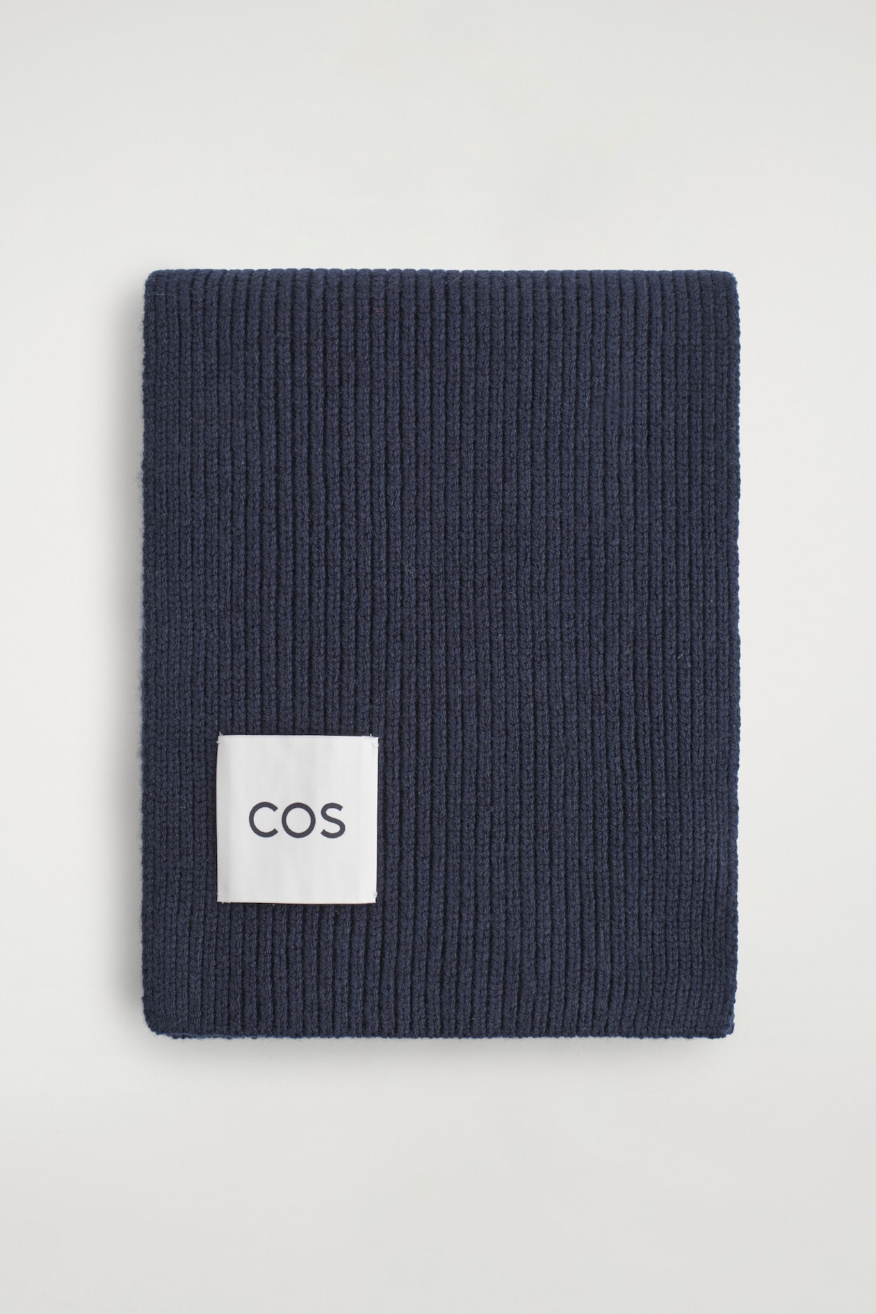 A navy blue ribbed knit scarf is folded flat and facing front, displaying its vertical ribbed texture. A square white fabric label featuring the black "COS" brand name is stitched onto the lower left corner.