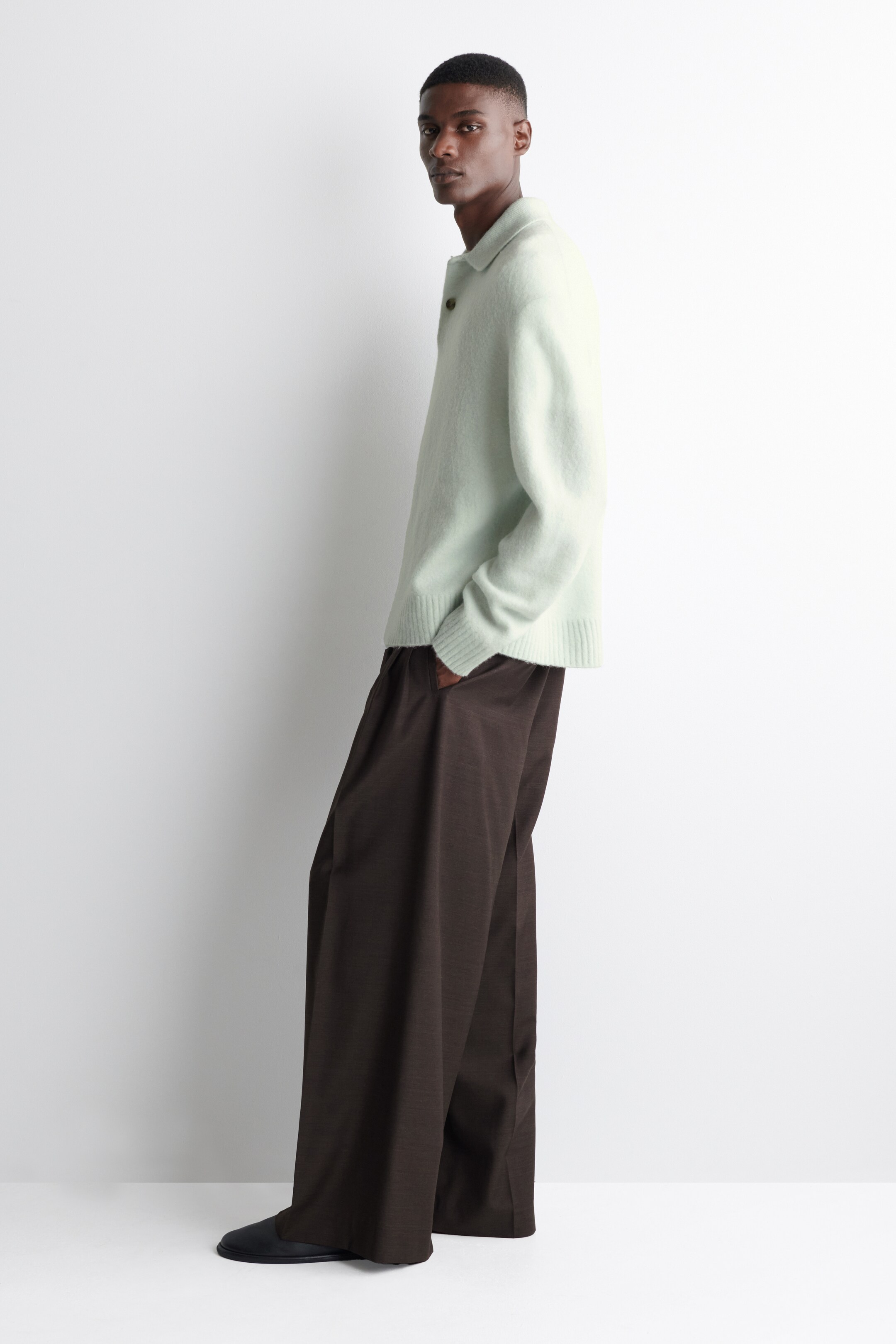 Visualizza immagine più grande: Un maglione polo verde chiaro viene indossato da una persona leggermente inclinata in avanti e rivolta a destra, mettendo in mostra la sua vestibilità rilassata, l'abbottonatura a due bottoni e i polsini e l'orlo a costine. Il maglione è abbinato a pantaloni a gamba larga marrone scuro che cadono liberamente sopra scarpe nere basse.