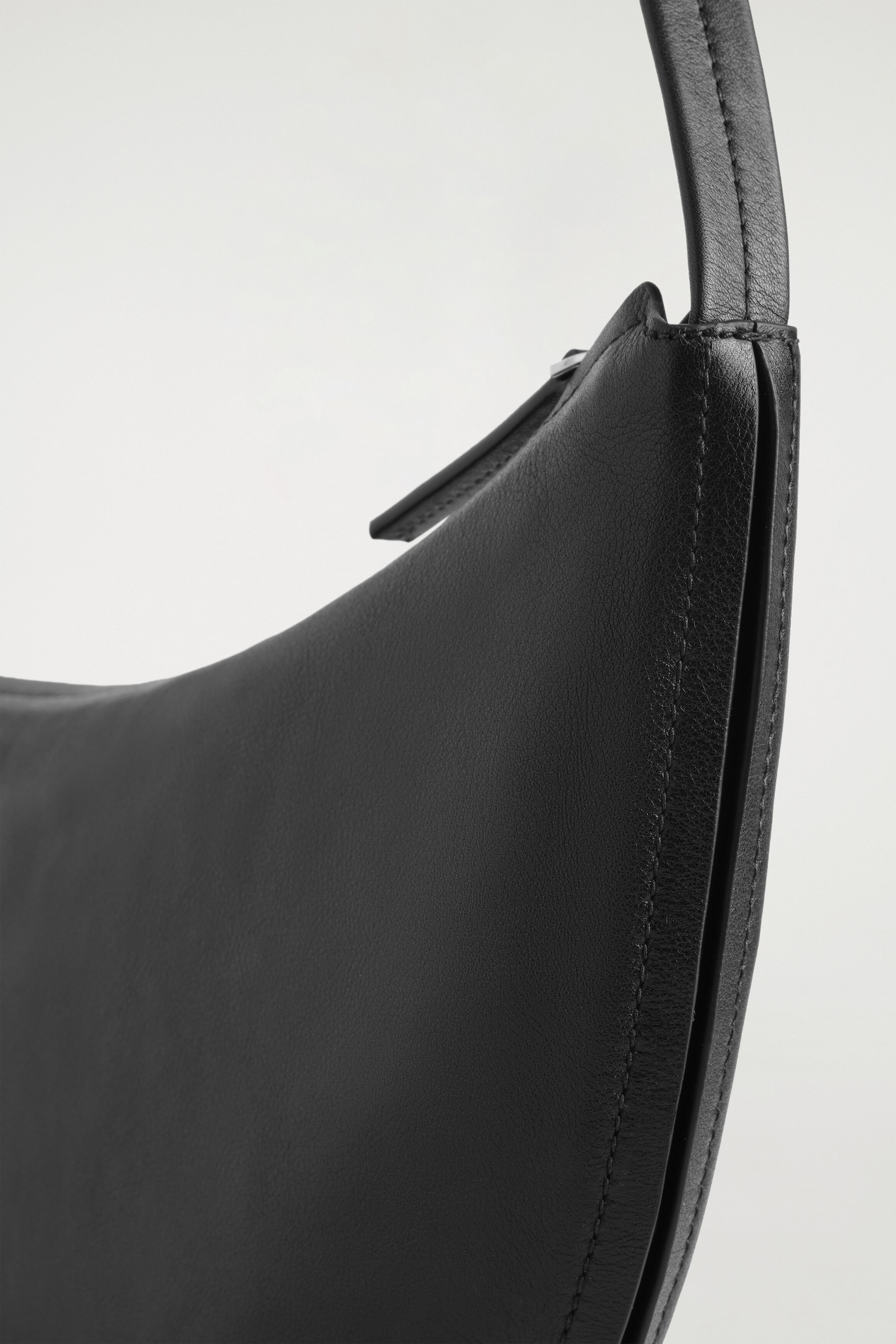 A black leather shoulder bag is shown in a close-up side view, highlighting its sculpted silhouette and soft-grain texture. The bag features a structured side panel with tonal stitching, a slim leather shoulder strap, and a zip closure with a leather pull tab.