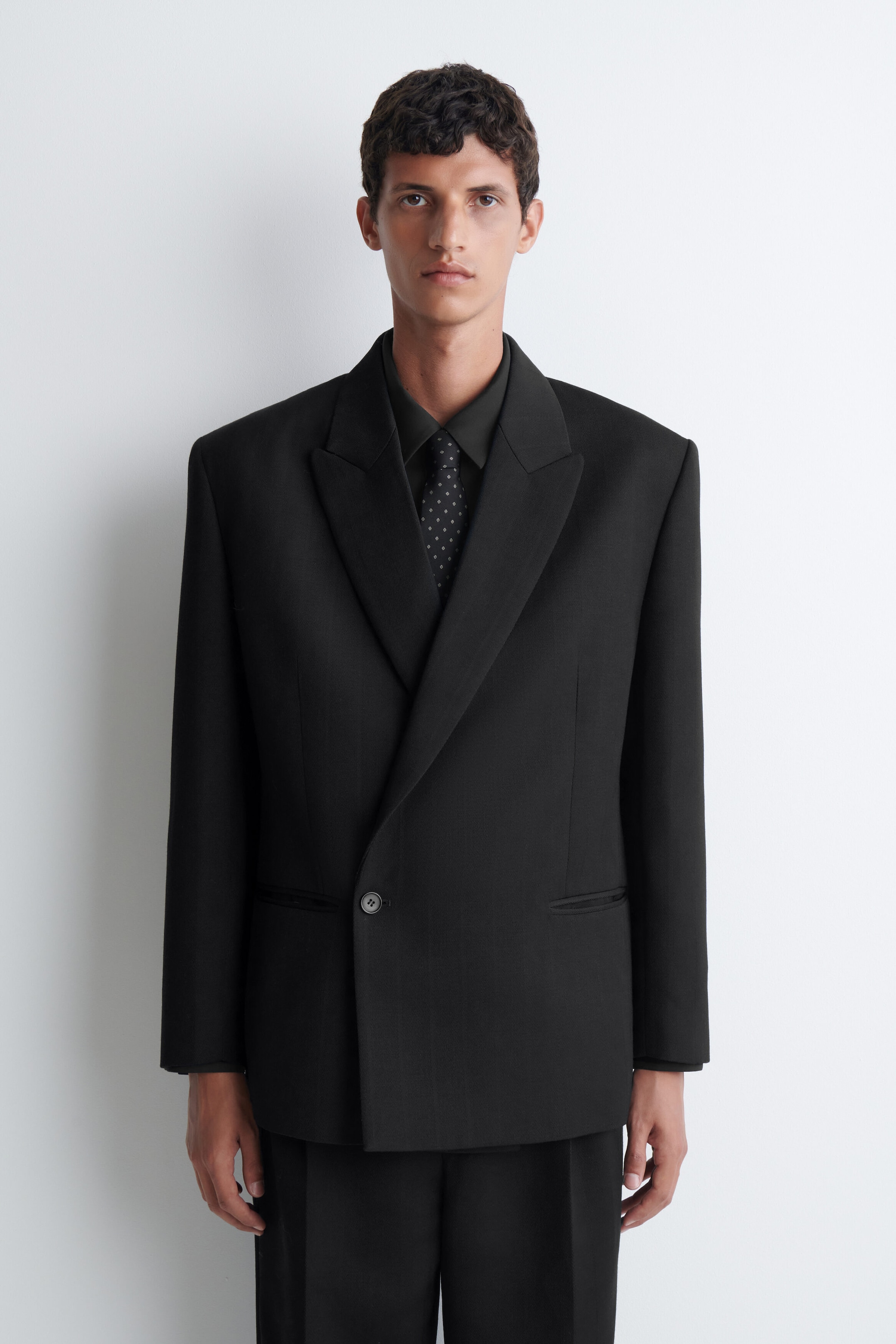 A black silk tie featuring a small, repeating foulard pattern is being worn by the model. The tie is layered under a black collared shirt and a black double-breasted blazer. A black silk tie featuring a small, repeating foulard pattern is being worn by the model. The tie is layered under a black collared shirt and a black double-breasted blazer.