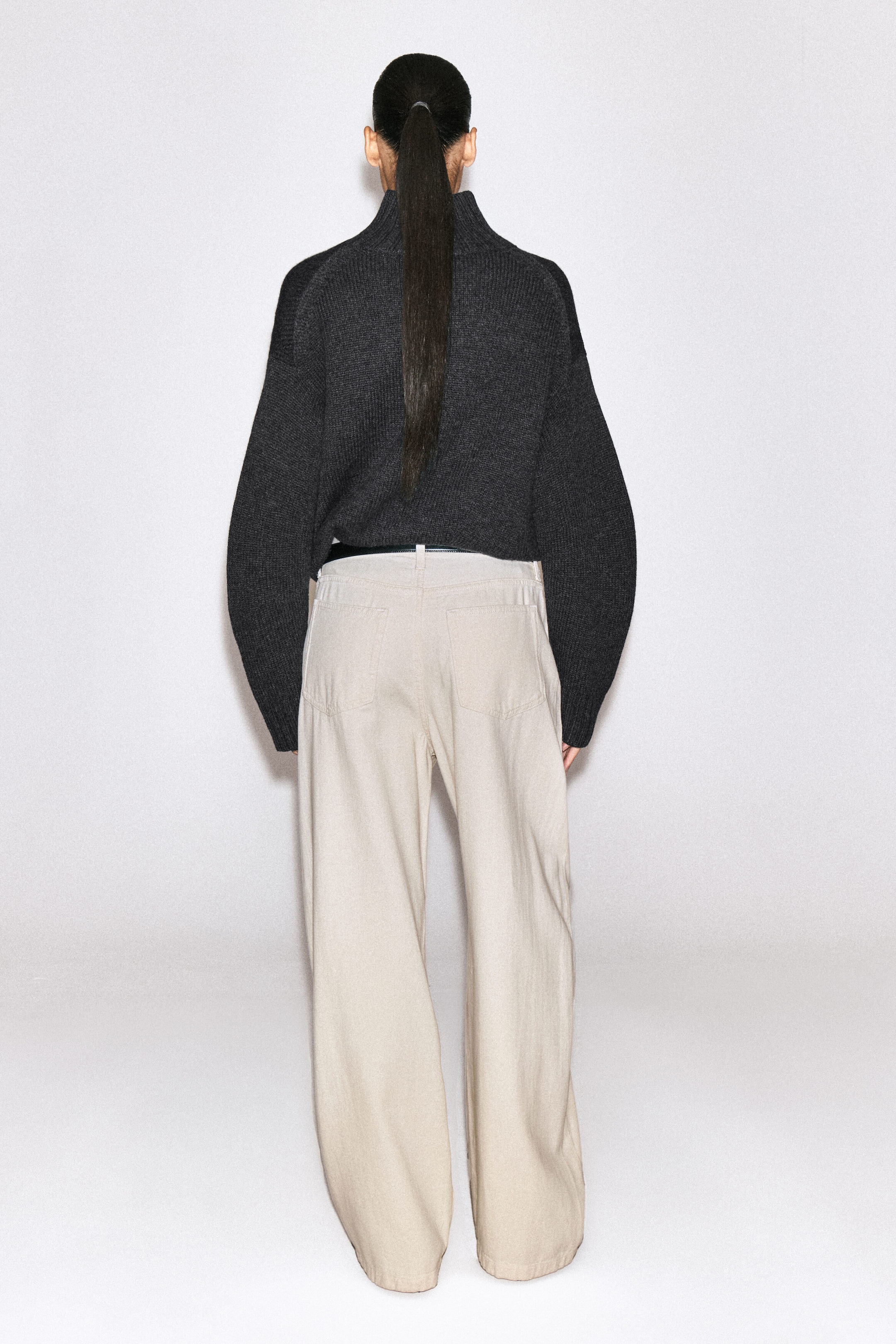 The Lagoon wide-leg jeans are shown from the back, showcasing a light beige denim fabric, a relaxed wide-leg silhouette, and two rear patch pockets. The jeans are styled with a thin black belt and a tucked-in dark grey knitted sweater. The Lagoon wide-leg jeans are shown from the back, showcasing a light beige denim fabric, a relaxed wide-leg silhouette, and two rear patch pockets. The jeans are styled with a thin black belt and a tucked-in dark grey knitted sweater.