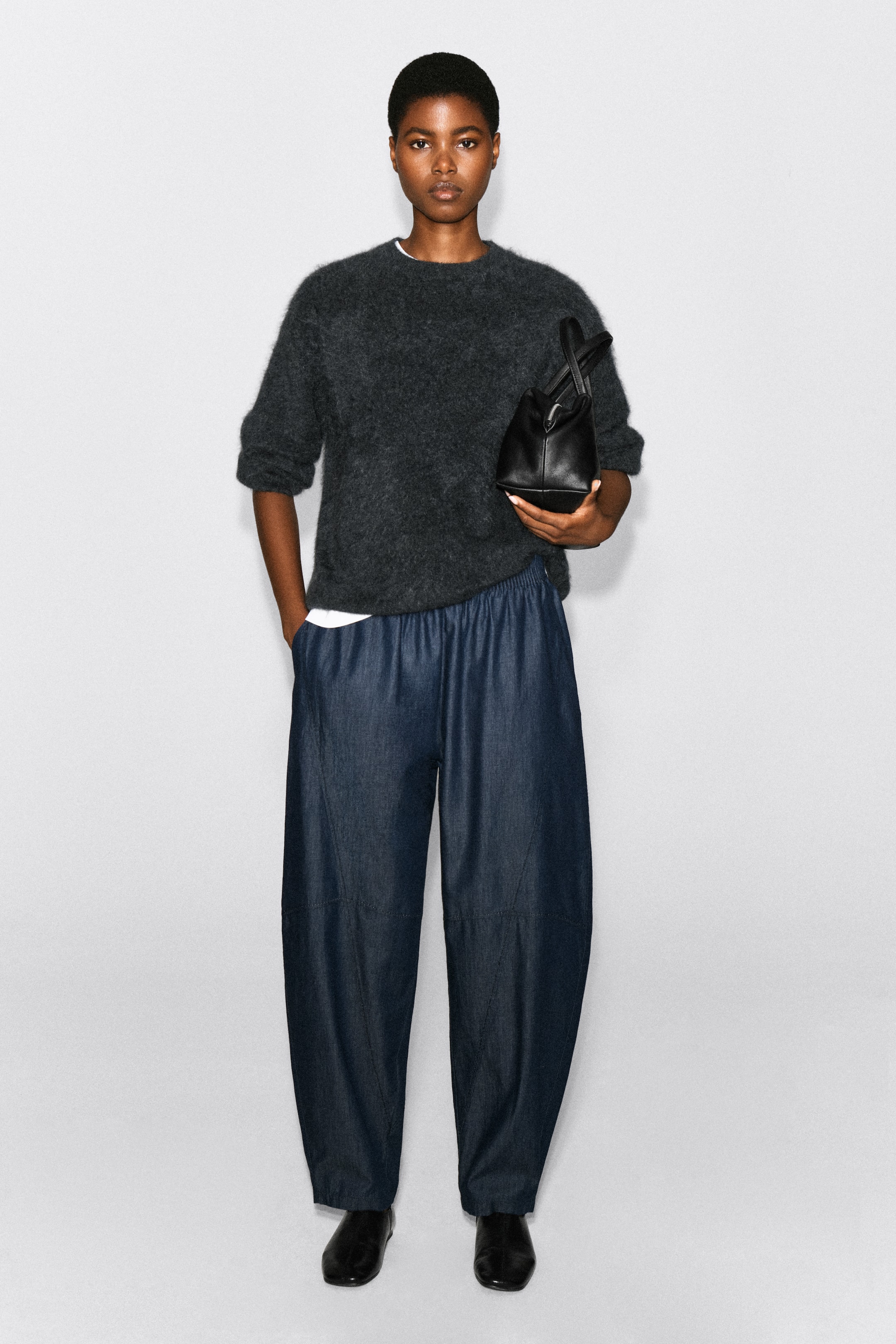 Visualizza l'immagine ingrandita: Pantaloni in denim elasticizzato a gamba a barile in lavaggio blu scuro indossati dalla modella, che è in piedi con una mano nella tasca laterale. I pantaloni dal taglio morbido, con gamba curva, sono abbinati a un maglione girocollo soffice grigio scuro e stivali neri in pelle.