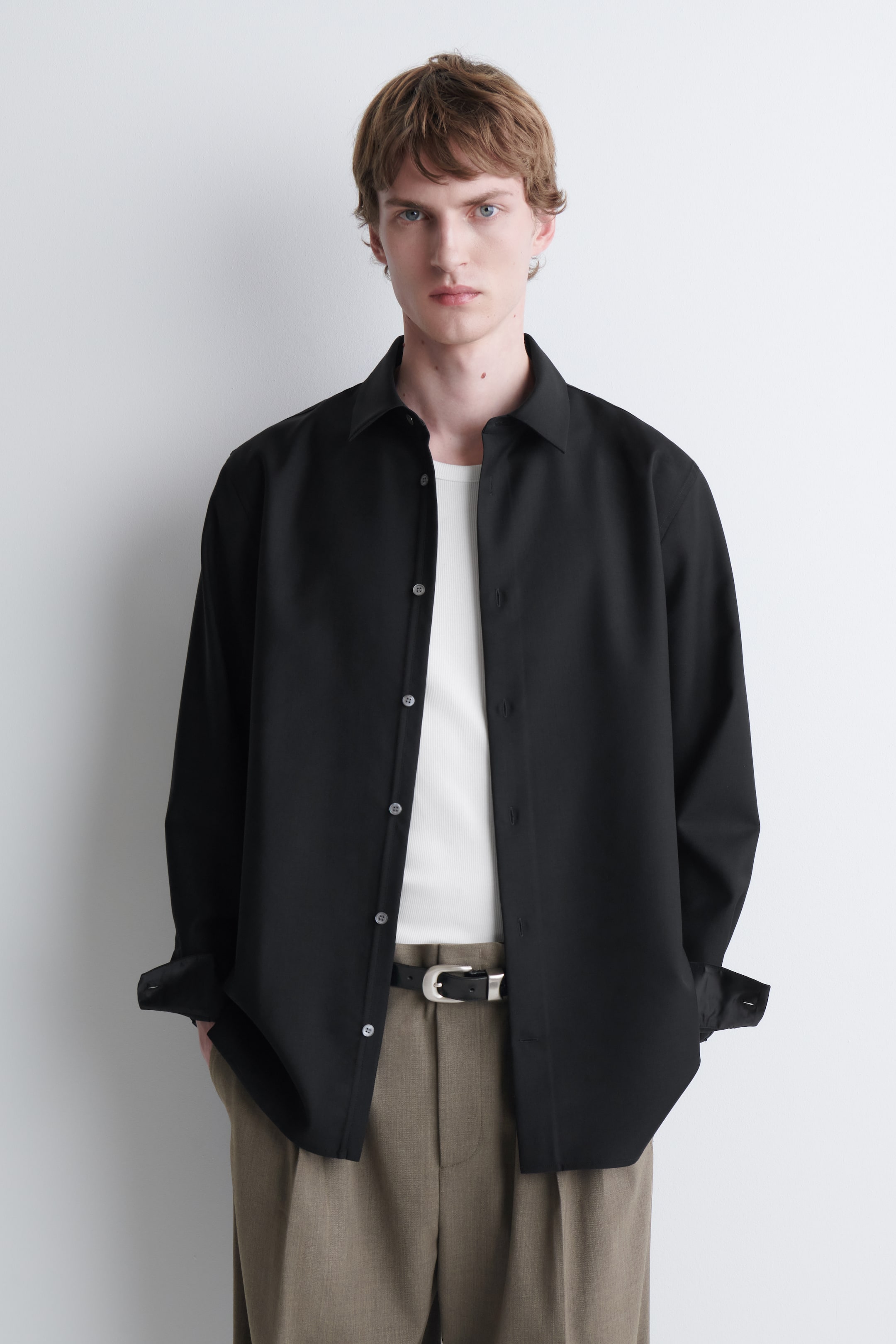 View larger image: A black relaxed wool-twill shirt is being worn open by the model, layered over a white T-shirt and styled with brown trousers. The shirt features a pointed collar, a button-down front, and long sleeves with undone, turned-back cuffs.