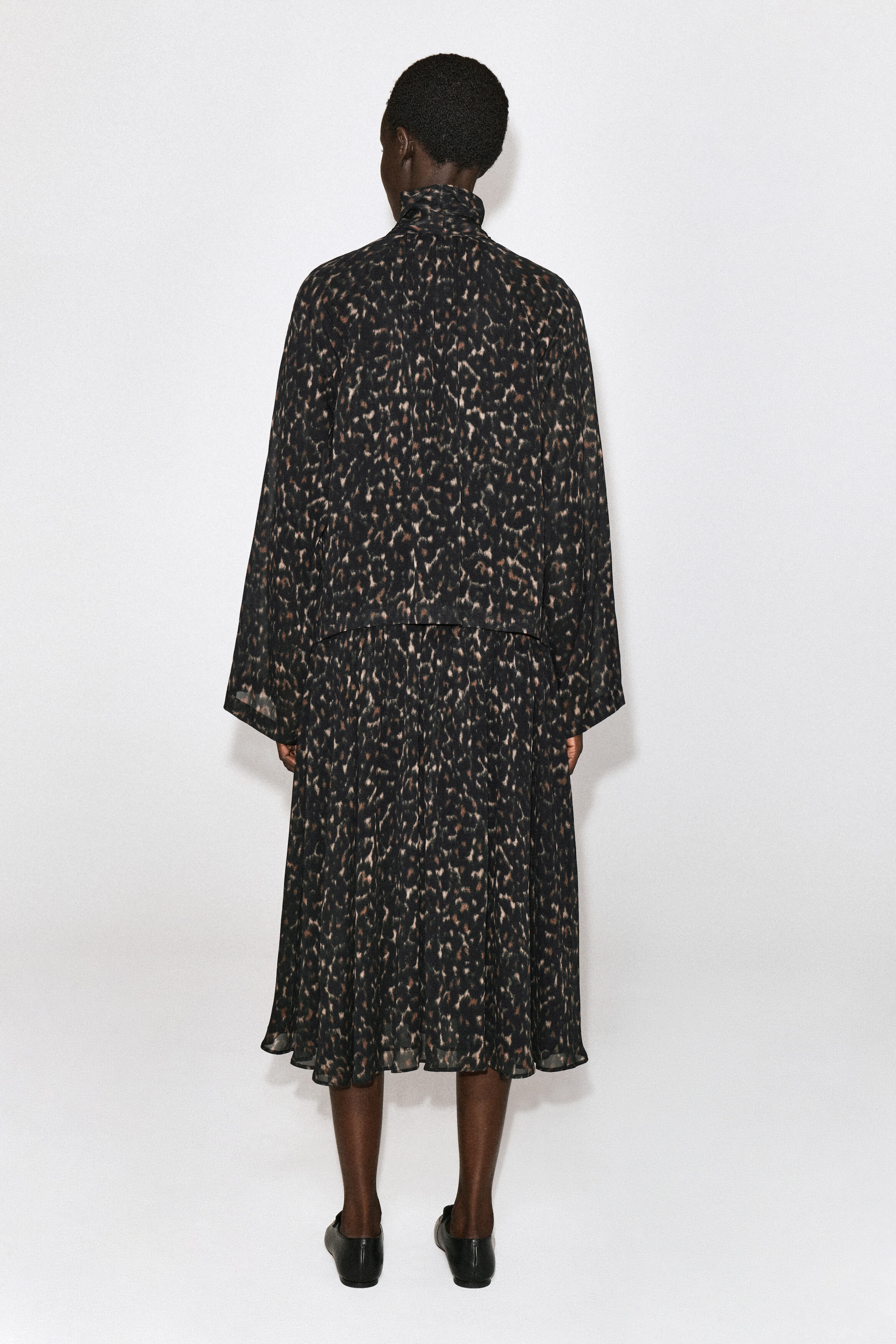 The leopard-print fluid scarf-detail dress is shown from the back, highlighting its high, wrapped collar and loose, wide long sleeves. The dress drapes in a layered silhouette, featuring a relaxed-fit top over a fluid, midi-length skirt. The leopard-print fluid scarf-detail dress is shown from the back, highlighting its high, wrapped collar and loose, wide long sleeves. The dress drapes in a layered silhouette, featuring a relaxed-fit top over a fluid, midi-length skirt.