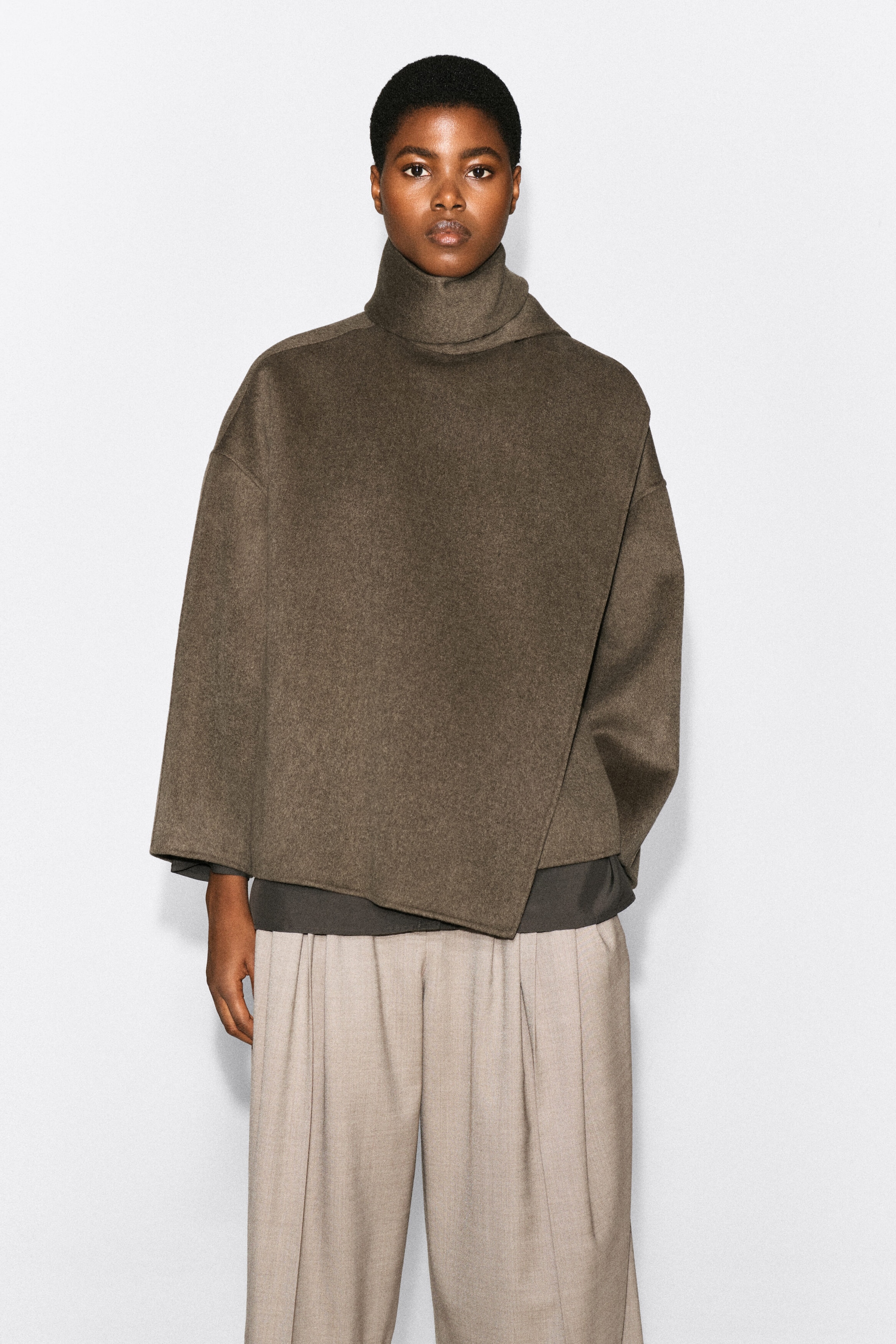 A double-faced wool-blend scarf jacket in a dark olive-brown colour is being worn by the model. The jacket features an integrated, wrapped scarf collar, a draped asymmetrical front, and wide, relaxed sleeves, styled over a dark grey underlayer and light beige pleated trousers. A double-faced wool-blend scarf jacket in a dark olive-brown colour is being worn by the model. The jacket features an integrated, wrapped scarf collar, a draped asymmetrical front, and wide, relaxed sleeves, styled over a dark grey underlayer and light beige pleated trousers.