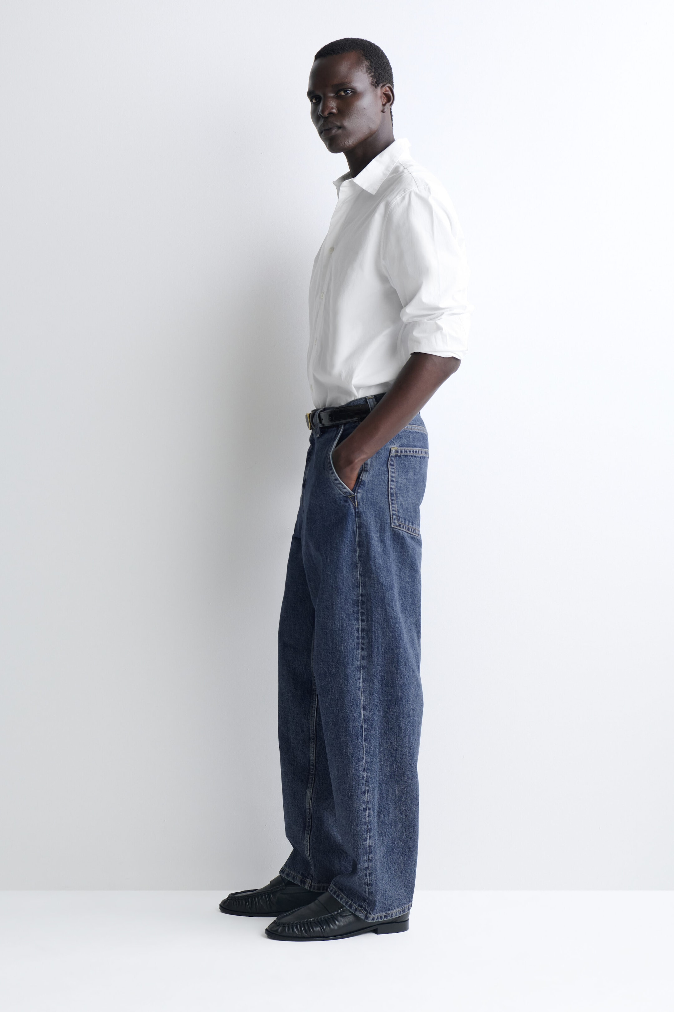 Relaxed-fit straight-leg denim trousers in a medium blue wash fall in a loose, straight-leg silhouette, shown from a side profile facing left. The trousers feature a classic five-pocket design and pool slightly over black leather loafers, styled with a tucked-in white shirt and a black belt. Relaxed-fit straight-leg denim trousers in a medium blue wash fall in a loose, straight-leg silhouette, shown from a side profile facing left. The trousers feature a classic five-pocket design and pool slightly over black leather loafers, styled with a tucked-in white shirt and a black belt.