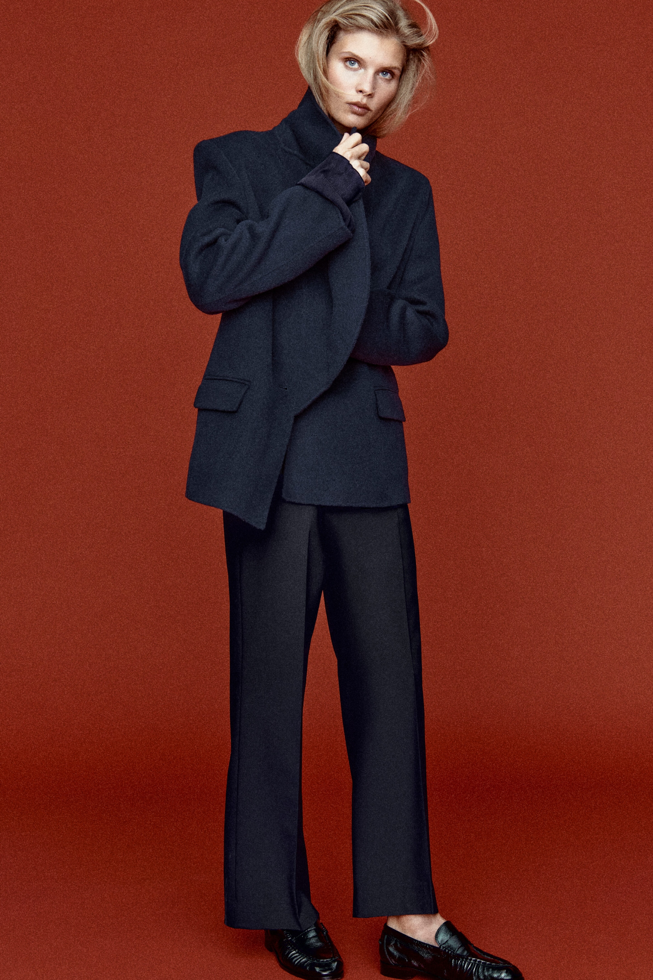 A model is facing front, wearing a dark navy tailored draped wool-blend blazer with structured shoulders, a high draped collar, and side flap pockets. She is pairing the blazer with matching dark navy twill cropped flared trousers featuring pressed centre creases and black leather loafers.