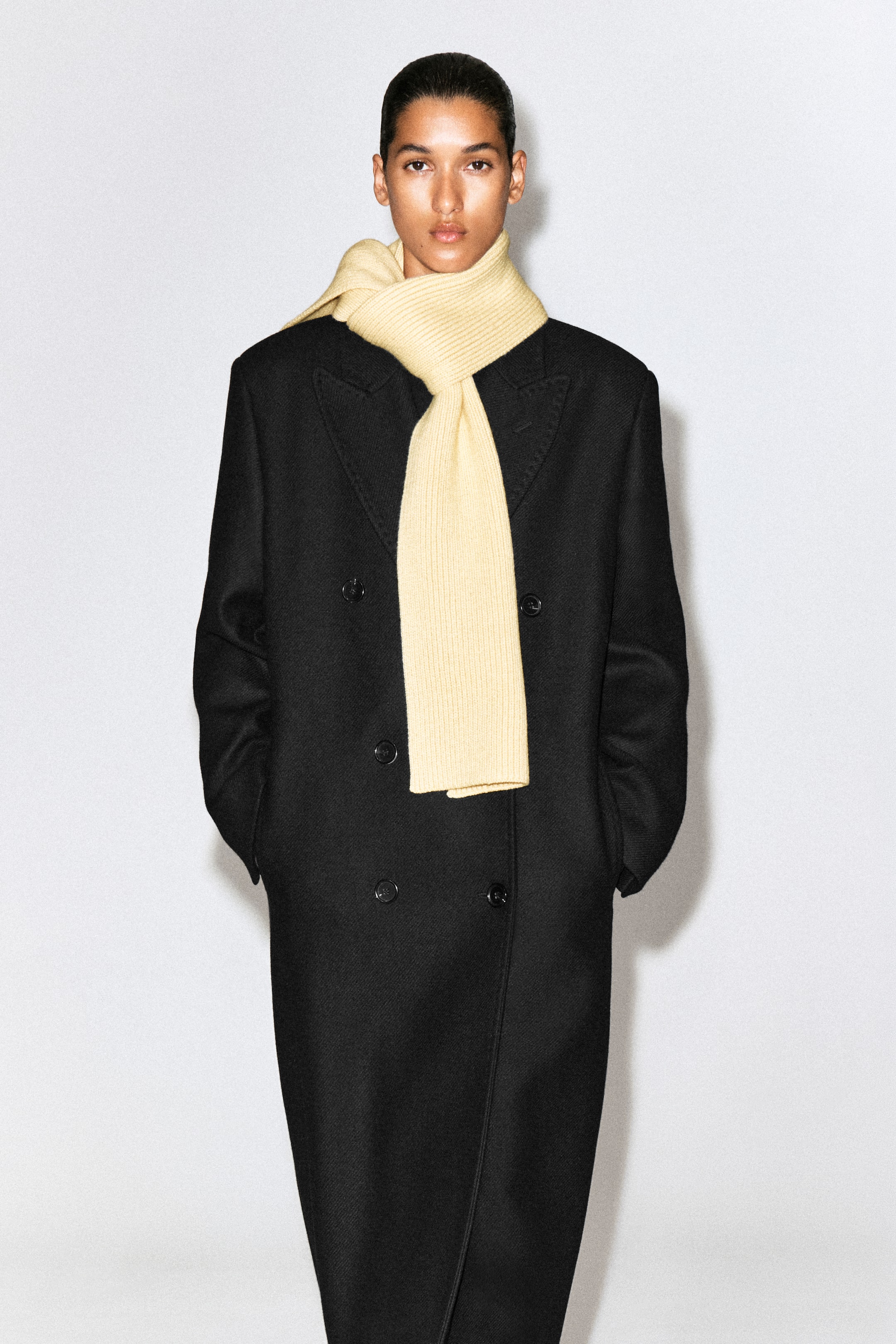 View larger image: The chunky ribbed wool scarf in pale yellow is shown from the front, wrapped around the neck of a model wearing a double-breasted black coat. The scarf features a textured ribbed knit construction and falls straight down the centre.