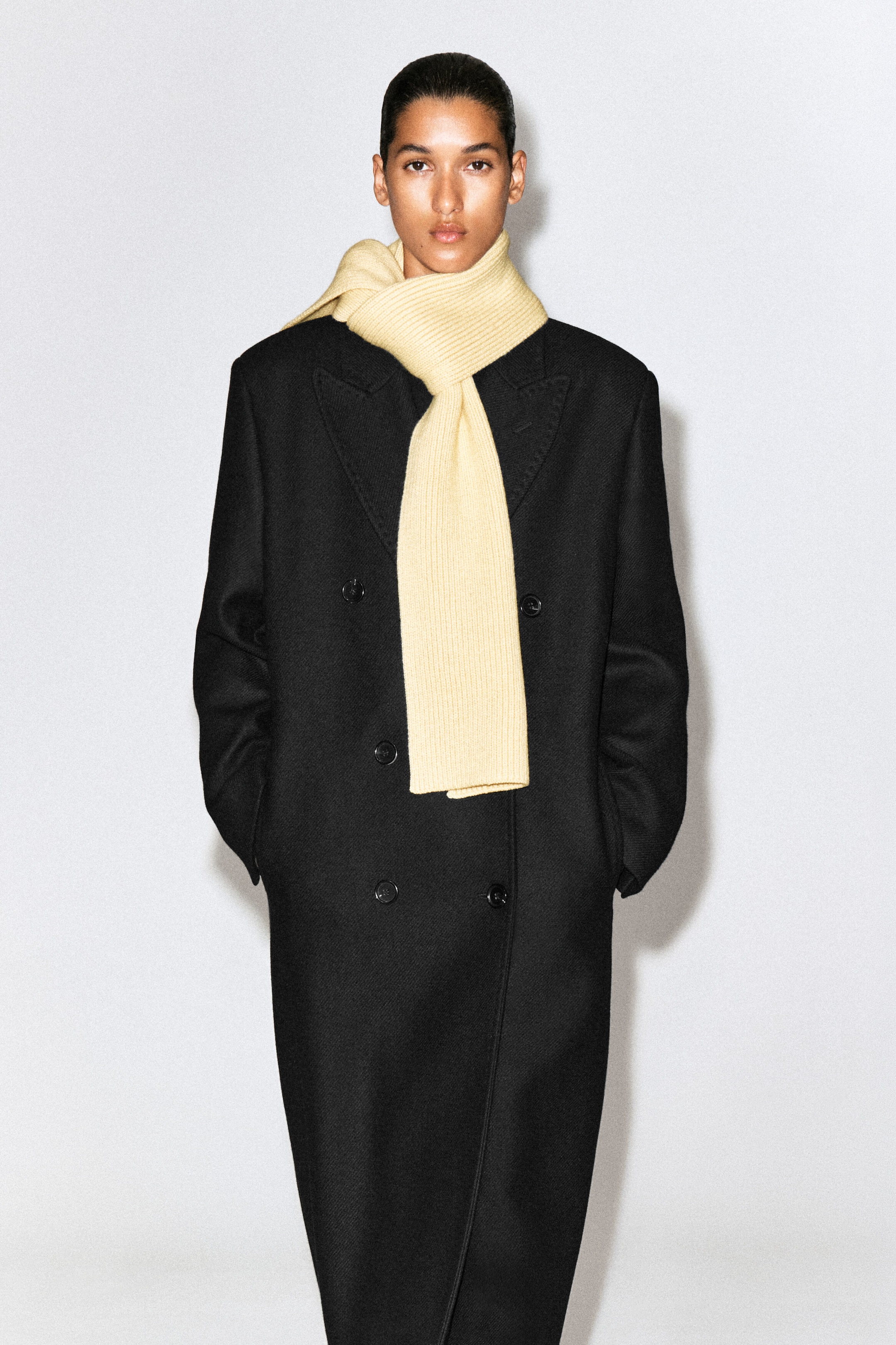 The chunky ribbed wool scarf in pale yellow is shown from the front, wrapped around the neck of a model wearing a double-breasted black coat. The scarf features a textured ribbed knit construction and drapes straight down the center. The chunky ribbed wool scarf in pale yellow is shown from the front, wrapped around the neck of a model wearing a double-breasted black coat. The scarf features a textured ribbed knit construction and drapes straight down the center.
