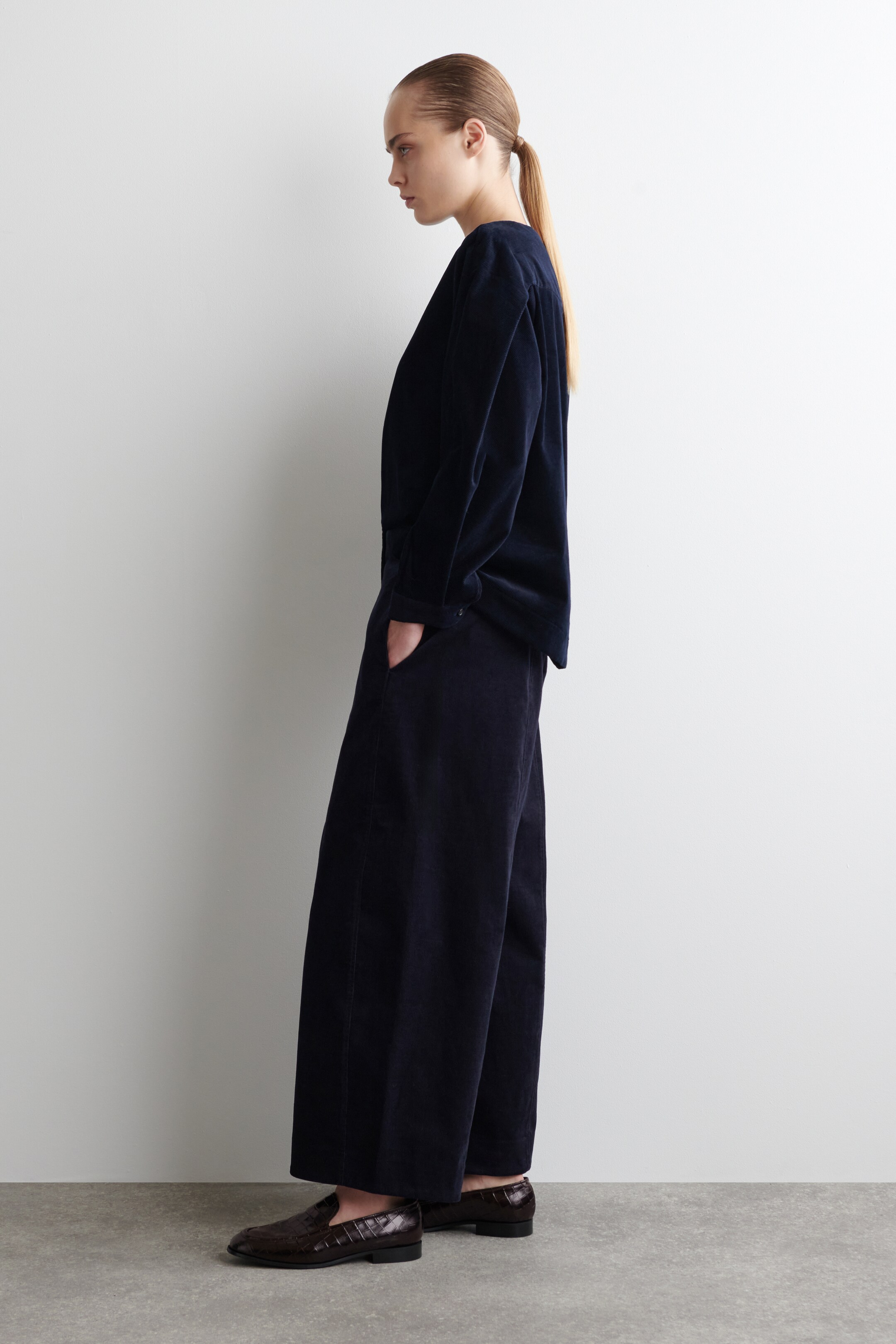 Ver imagen más grande: Un conjunto de dos piezas de pana azul oscuro se presenta desde una vista lateral, con una blusa de corte holgado de manga larga y pantalones de pierna ancha a juego. Los pantalones incluyen bolsillos laterales, con una mano descansando dentro, y están combinados con mocasines de cuero grabado en marrón oscuro.