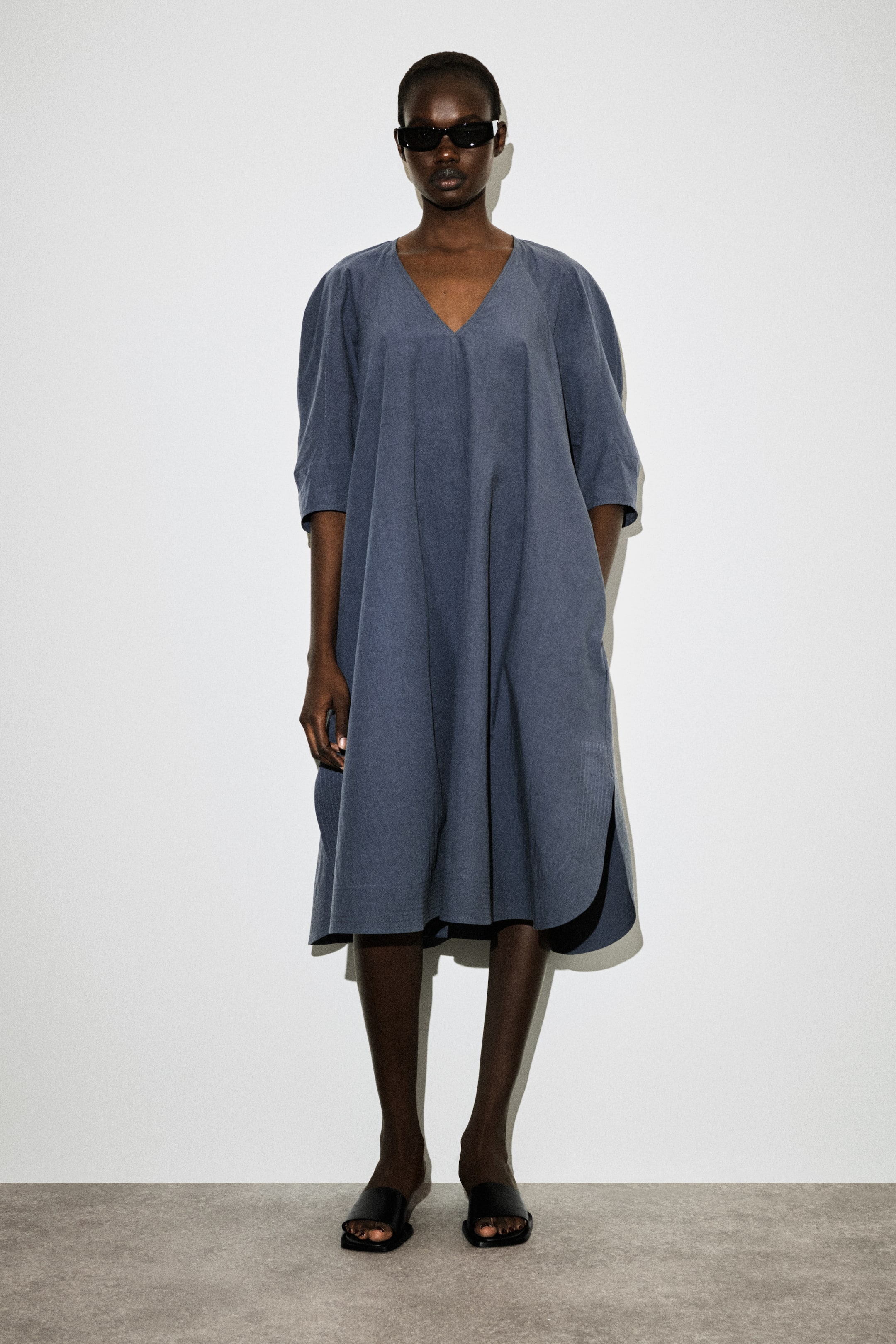 הצגת תמונה גדולה יותר: שמלת המידי בצבע כחול-אפור מתאפיינת במחשוף בצורת V, שרוולים נפוחים באורך שלושה רבעים וחריצים בצדדים בשולי השמלה, ומציגה סילואט A-ליין נינוח בבד בעל מרקם.