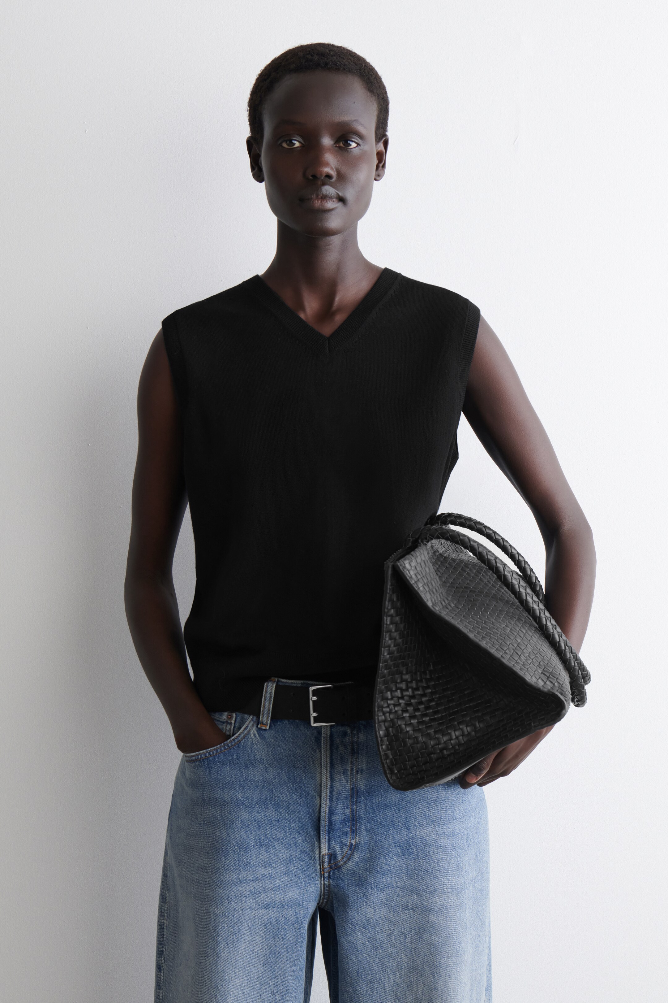 Visualizza immagine più grande: Un top nero senza maniche in maglia, con scollo a V e orlo dritto, viene presentato frontalmente. Una borsa in pelle intrecciata nera con un manico spesso intrecciato viene tenuta in mano, mostrando la sua trama intricata e la forma morbida e floscia.