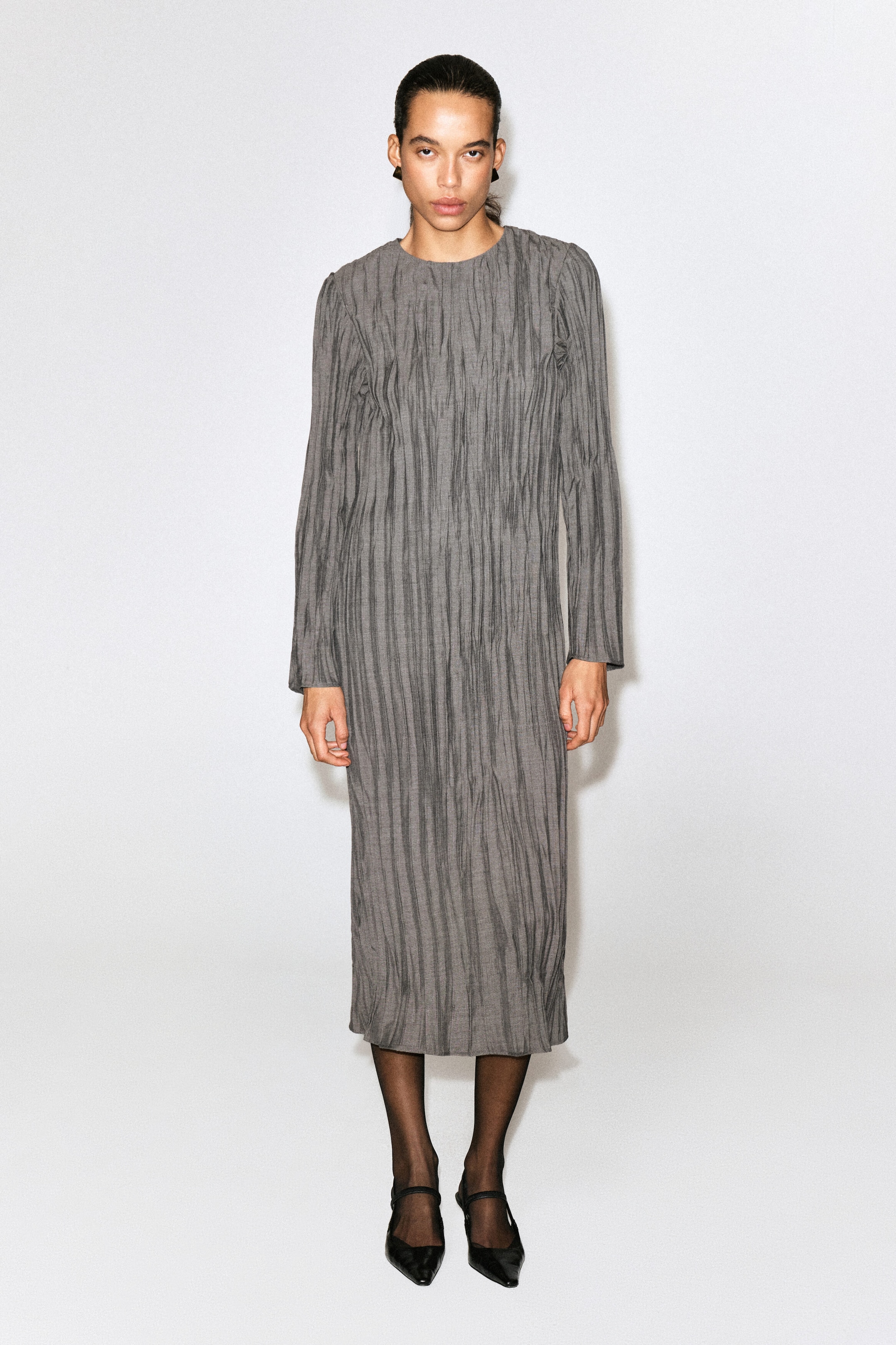This gray midi dress is shown from the front, featuring a vertically crinkled texture, a crew neck, and long sleeves. It drapes in a relaxed, straight silhouette that falls to a midi length.