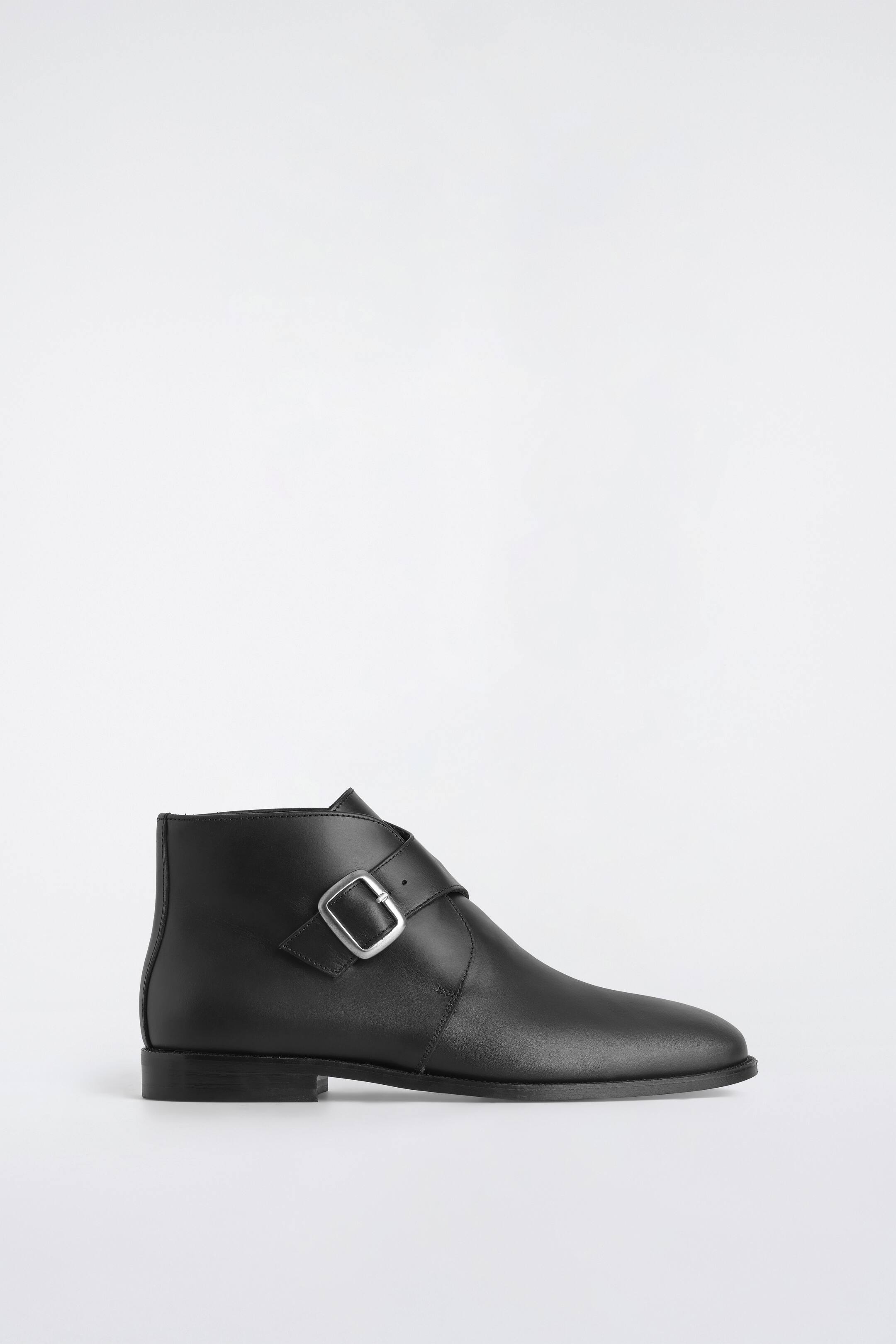 Grotere afbeelding bekijken: Een zwarte leren monk boot is in zijaanzicht naar rechts gericht weergegeven, met een gladde matte leren afwerking en een enkelhoge silhouet. Een brede diagonale band loopt over de voorkant van de enkel en wordt vastgemaakt met een zilverkleurige metalen gesp.