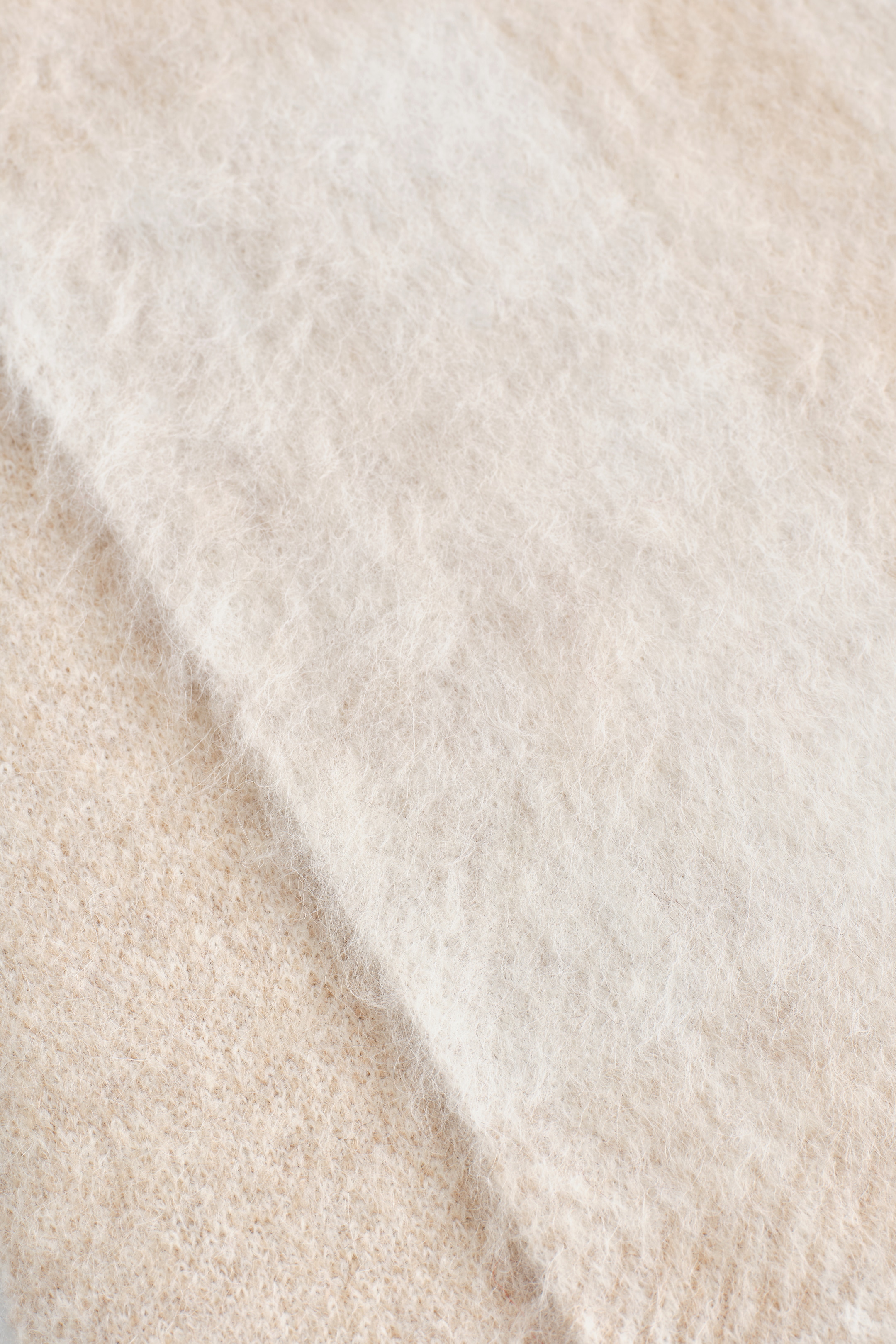 Agrandir l'image: Une écharpe met en avant un gros plan de son matériau, avec une section de fibres couleur crème clair, fortement brossées, à côté d'une section de tissu tricoté finement, de couleur beige clair.