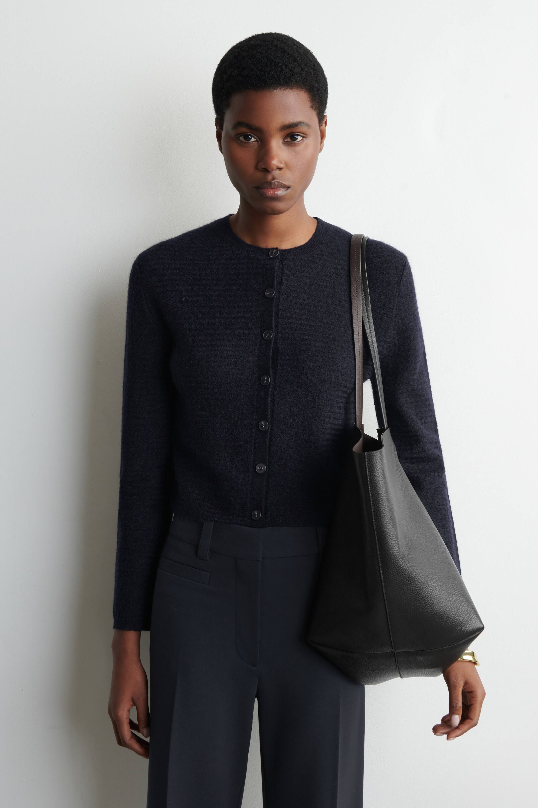 הצגת תמונה גדולה יותר: הדוגמנית לובשת קרדיגן בגוון כהה, קצר, עם סגירת כפתורים וטקסטורה מצולעת. תיק יד מעור שחור עם רצועות רחבות נישא על כתפה של הדוגמנית.