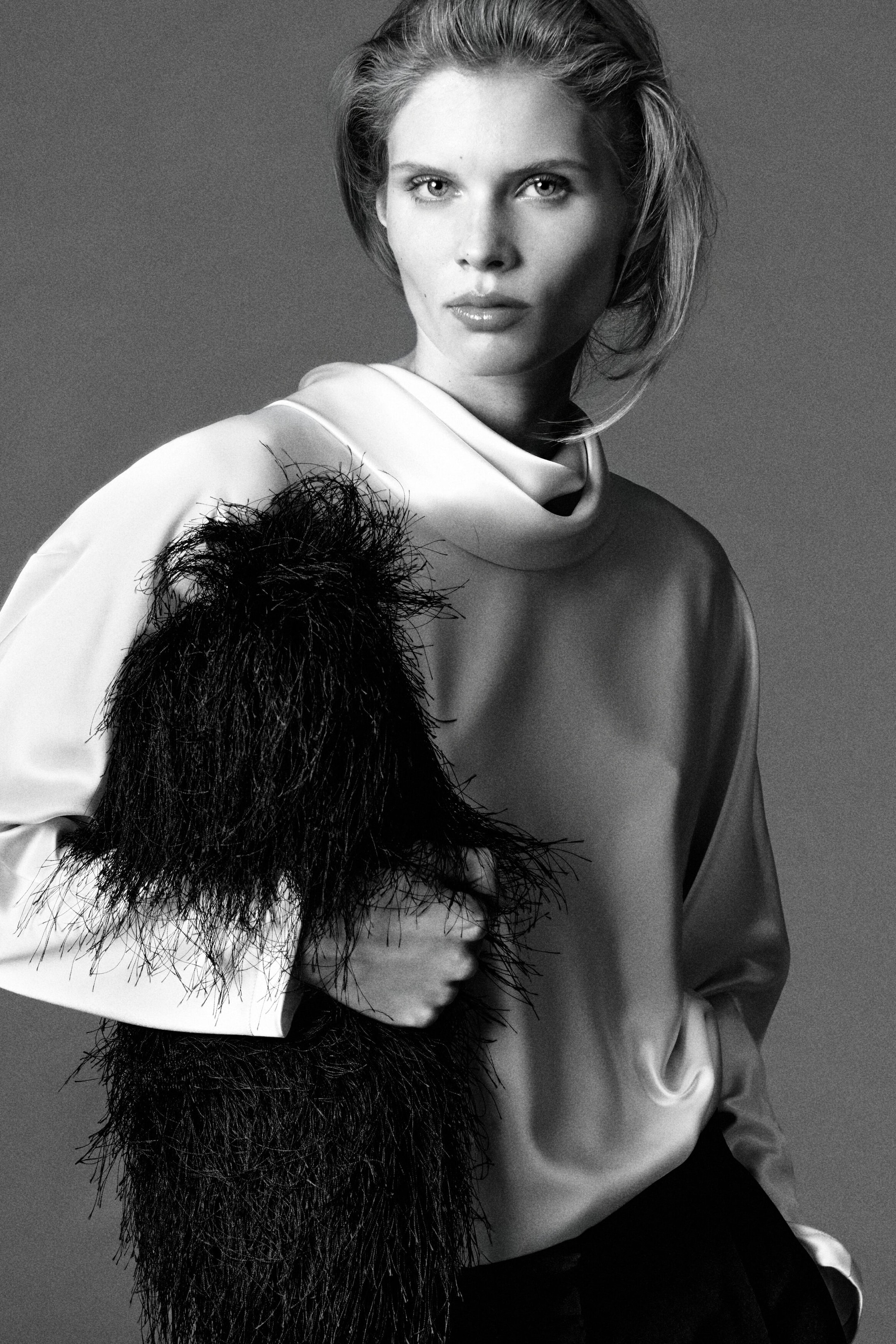 A front-facing model is wearing a light-colored cowl-neck satin blouse with a draped neckline and long sleeves, paired with dark pleated silk-crepe barrel-leg pants visible at the waist. The model is holding a heavily textured black fringed clutch bag across the front of the body. A front-facing model is wearing a light-colored cowl-neck satin blouse with a draped neckline and long sleeves, paired with dark pleated silk-crepe barrel-leg pants visible at the waist. The model is holding a heavily textured black fringed clutch bag across the front of the body.