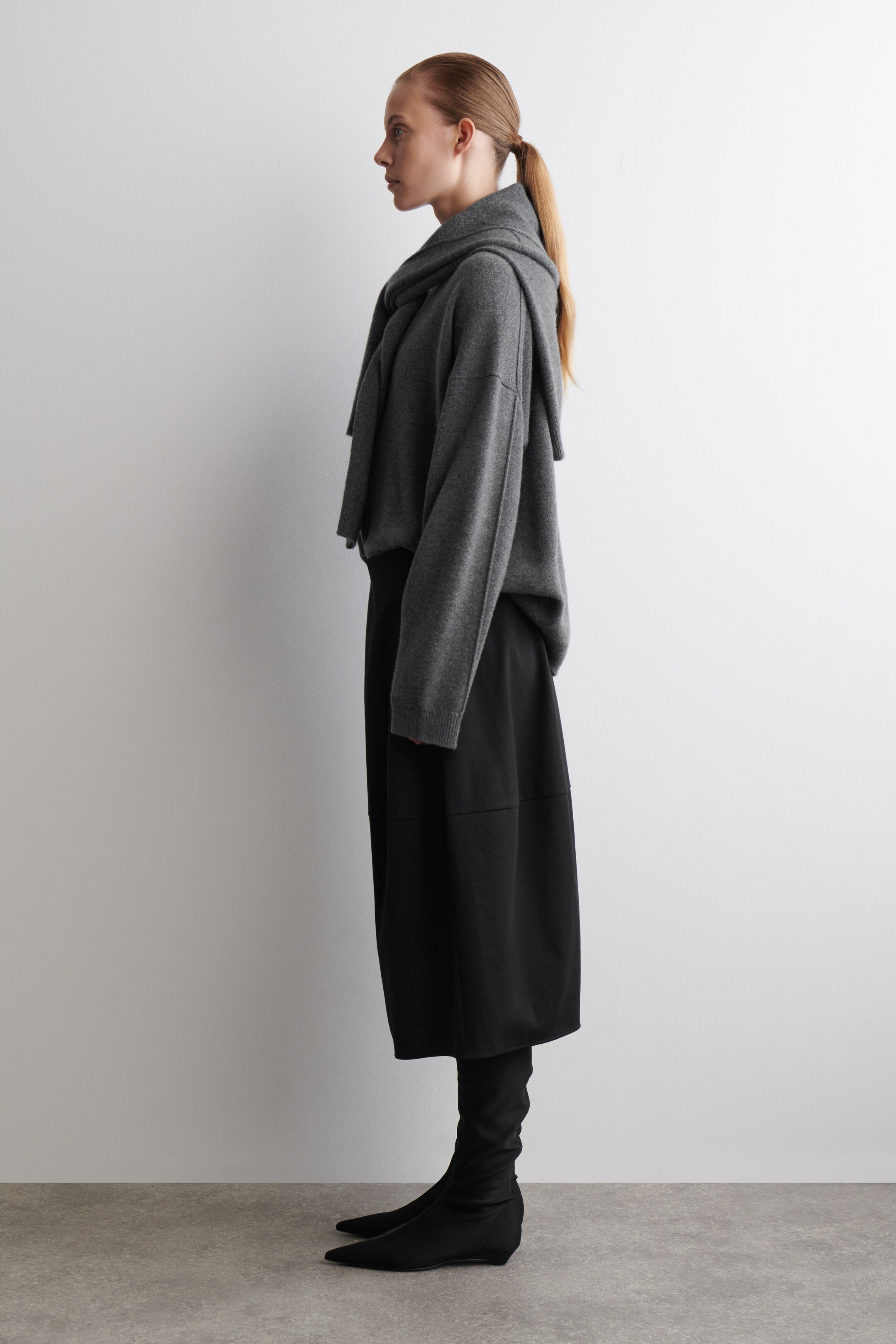 Agrandir l'image: Un pull en tricot gris, une jupe noire de longueur midi et des bottes-chaussettes noires sont présentés de profil. Le pull gris présente des coutures apparentes, un col drapé haut et des manches longues, superposant la jupe noire avec un panneau subtil, et des bottes-chaussettes noires à bout pointu qui s'étendent jusqu'au mollet.
