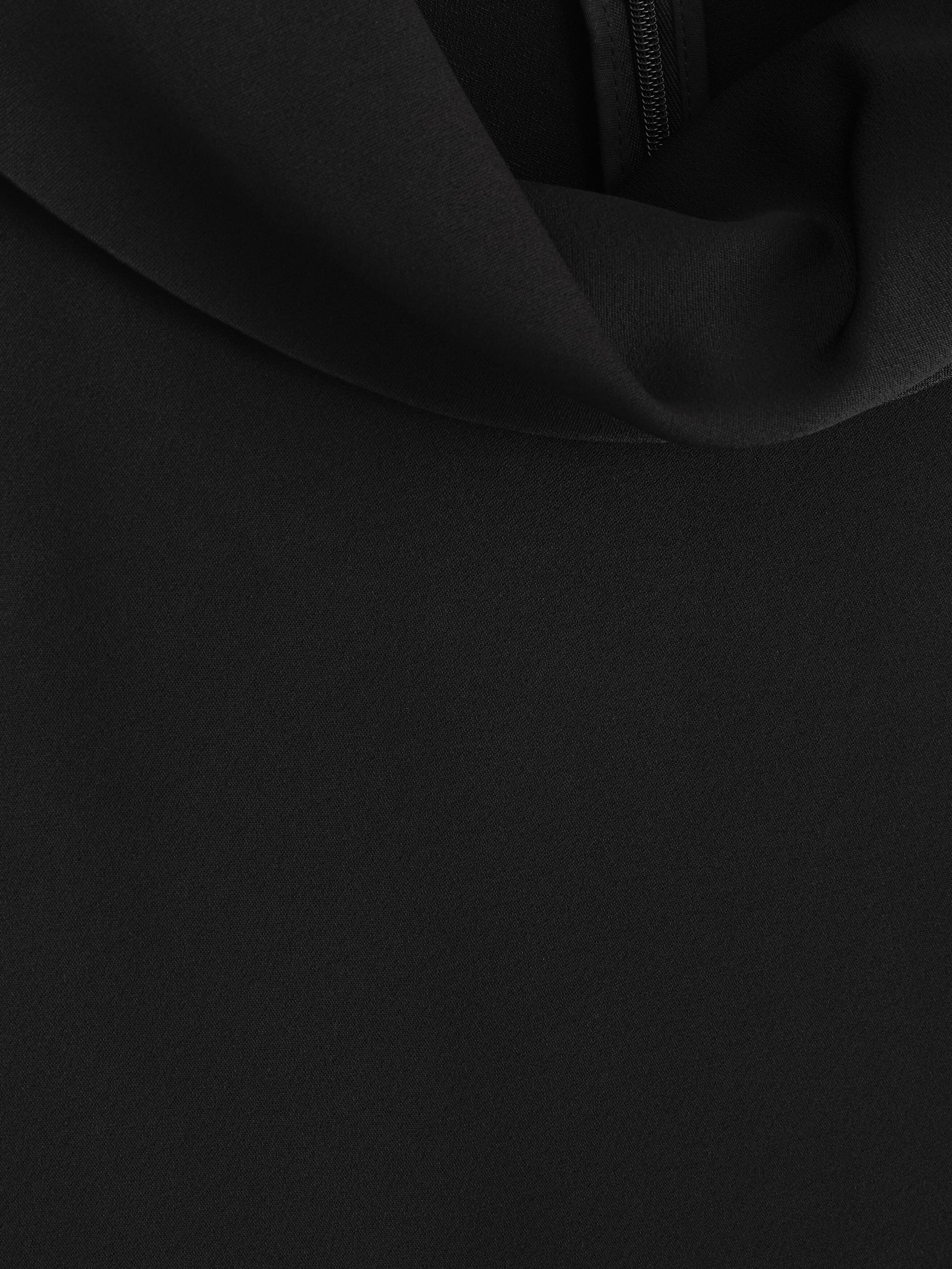 A close-up of the black crepe midi dress showing the finely textured matte fabric and the soft drape of the cowl neckline. A black zip is partially visible within the upper fold of the fabric.