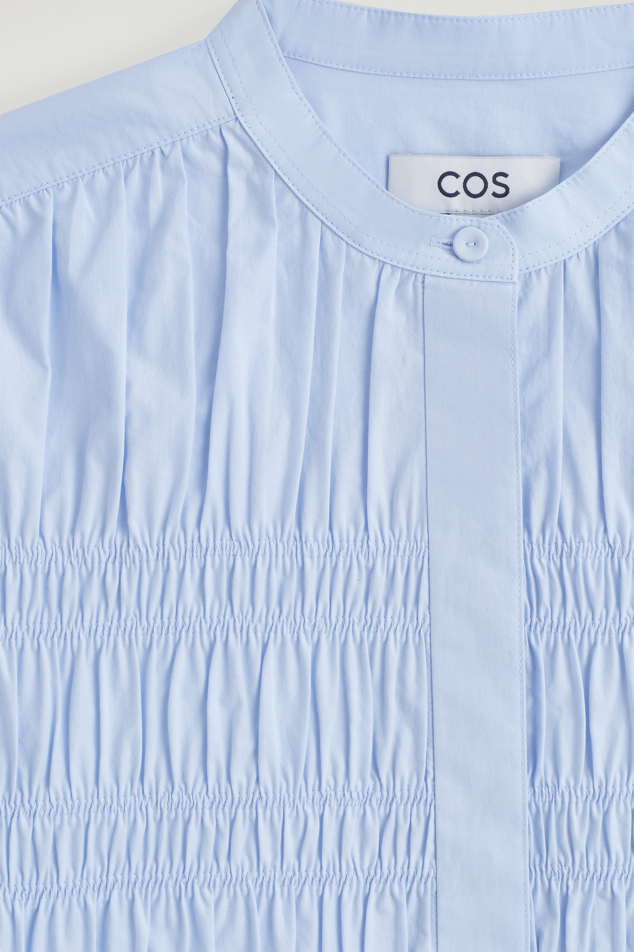 The ruched cotton blouse is facing front in a close-up view, showcasing a band collar with a single top button and a concealed placket. The bodice is displaying dense smocked ruching that is creating vertical gathers across the light blue fabric. The ruched cotton blouse is facing front in a close-up view, showcasing a band collar with a single top button and a concealed placket. The bodice is displaying dense smocked ruching that is creating vertical gathers across the light blue fabric.