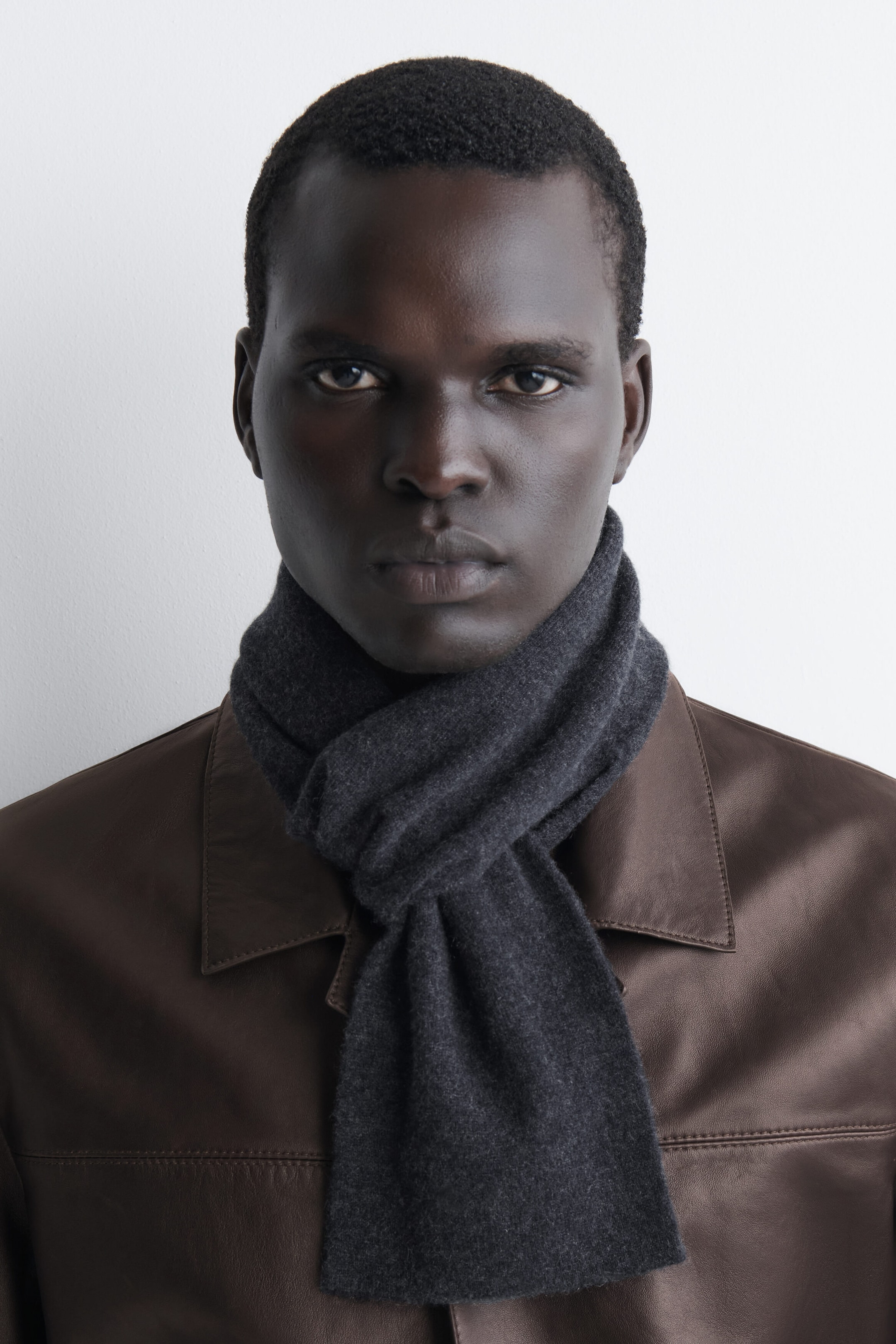 A dark gray cashmere pull-through scarf wraps snugly around the neck in a front-facing view, with one end pulled through the other and draping straight down over a brown leather jacket. A dark gray cashmere pull-through scarf wraps snugly around the neck in a front-facing view, with one end pulled through the other and draping straight down over a brown leather jacket.