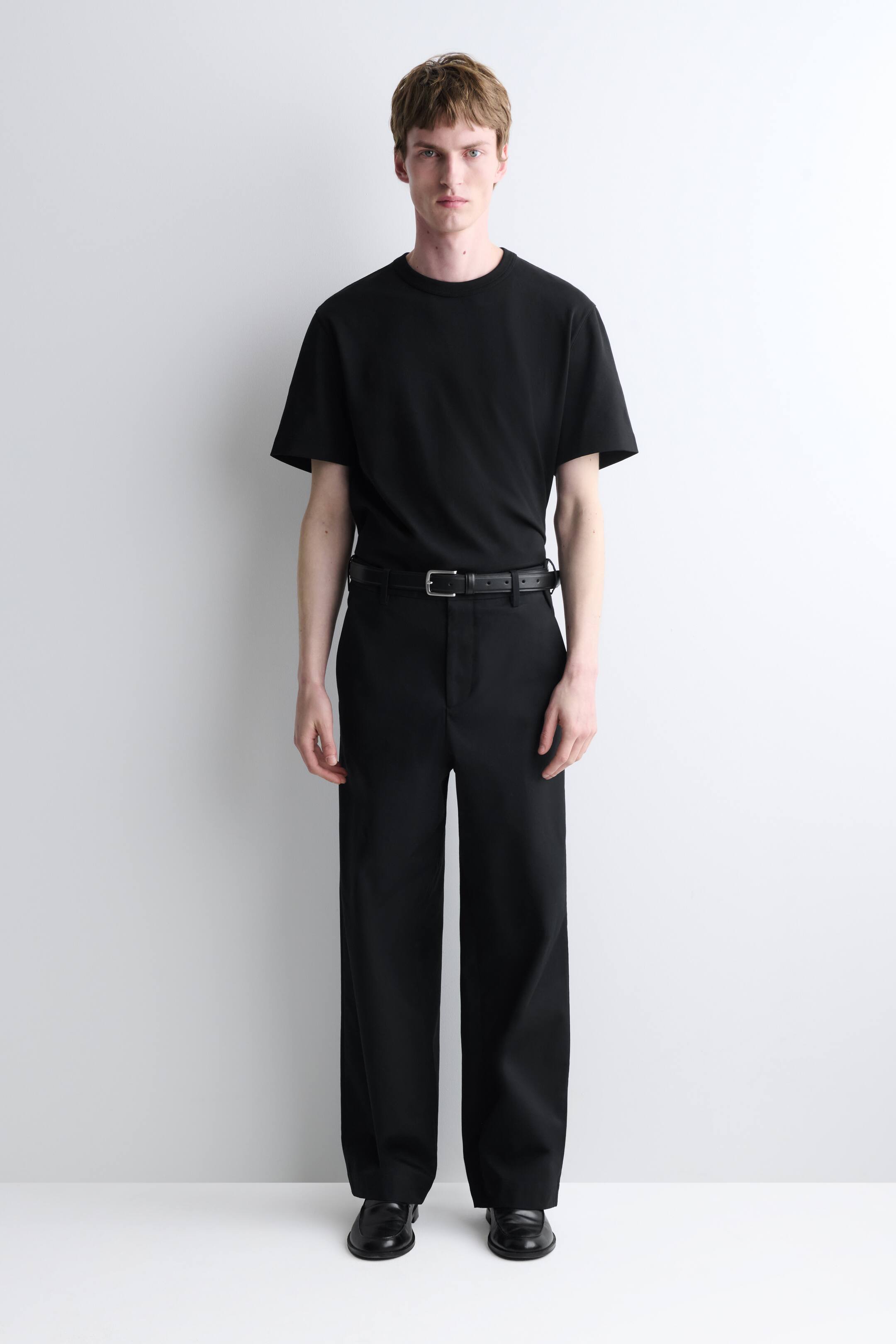 הצגת תמונה גדולה יותר: חולצת טי שחורה עם שרוולים קצרים וצווארון עגול נלבשת בתוך מכנסיים רחבים שחורים, המוחזקים על ידי חגורה שחורה עם אבזם בגוון כסף. כל התלבושת, כולל נעלי לופרס מעור שחור, מופנית קדימה, ומציגה את ההתאמה הכללית שלה.