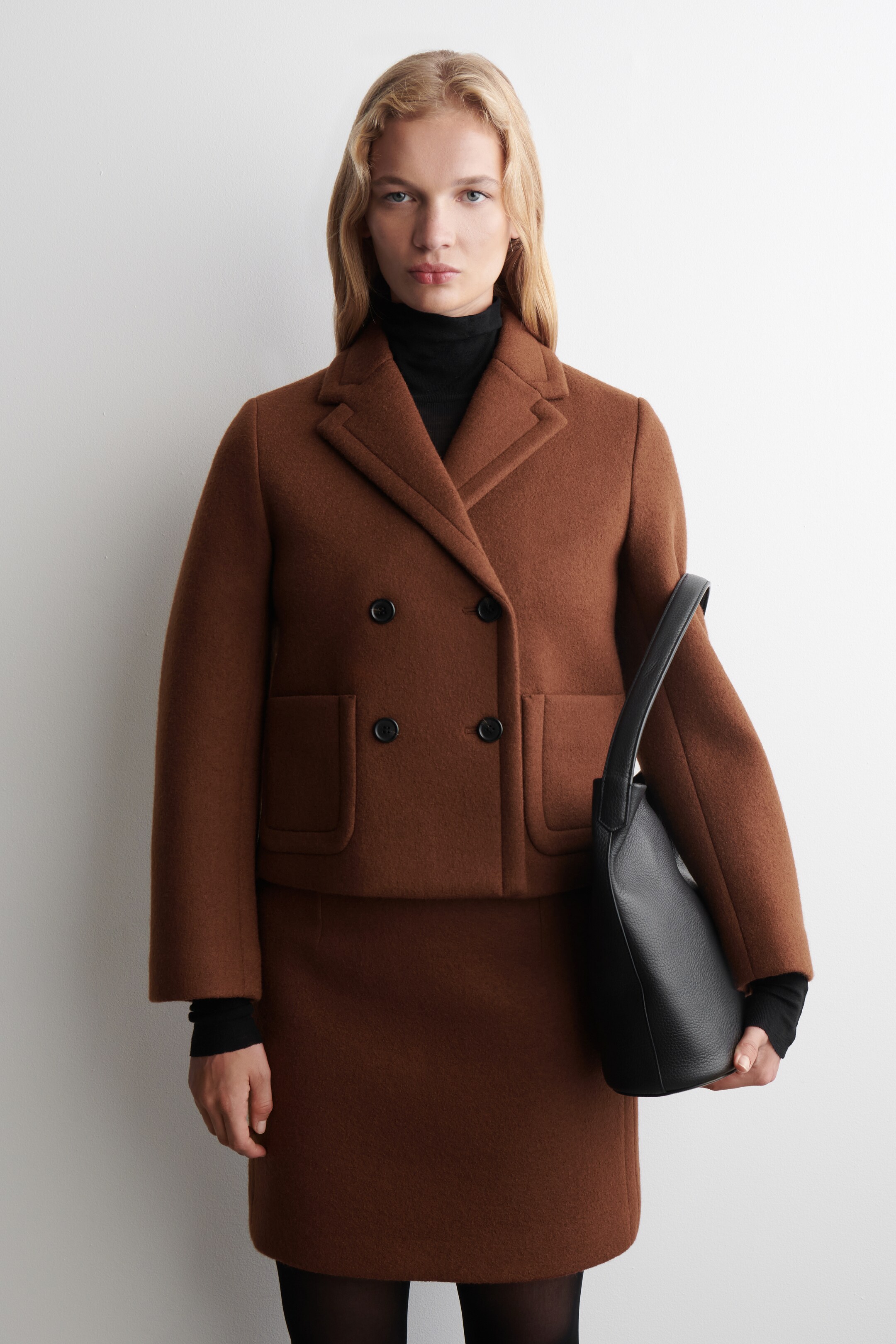View larger image: A brown double-breasted jacket is being shown from the front, displaying notch lapels, four black buttons, and two rounded patch pockets, while being worn over a black turtleneck and coordinating brown mini skirt. A black textured leather handbag is being held by the side.