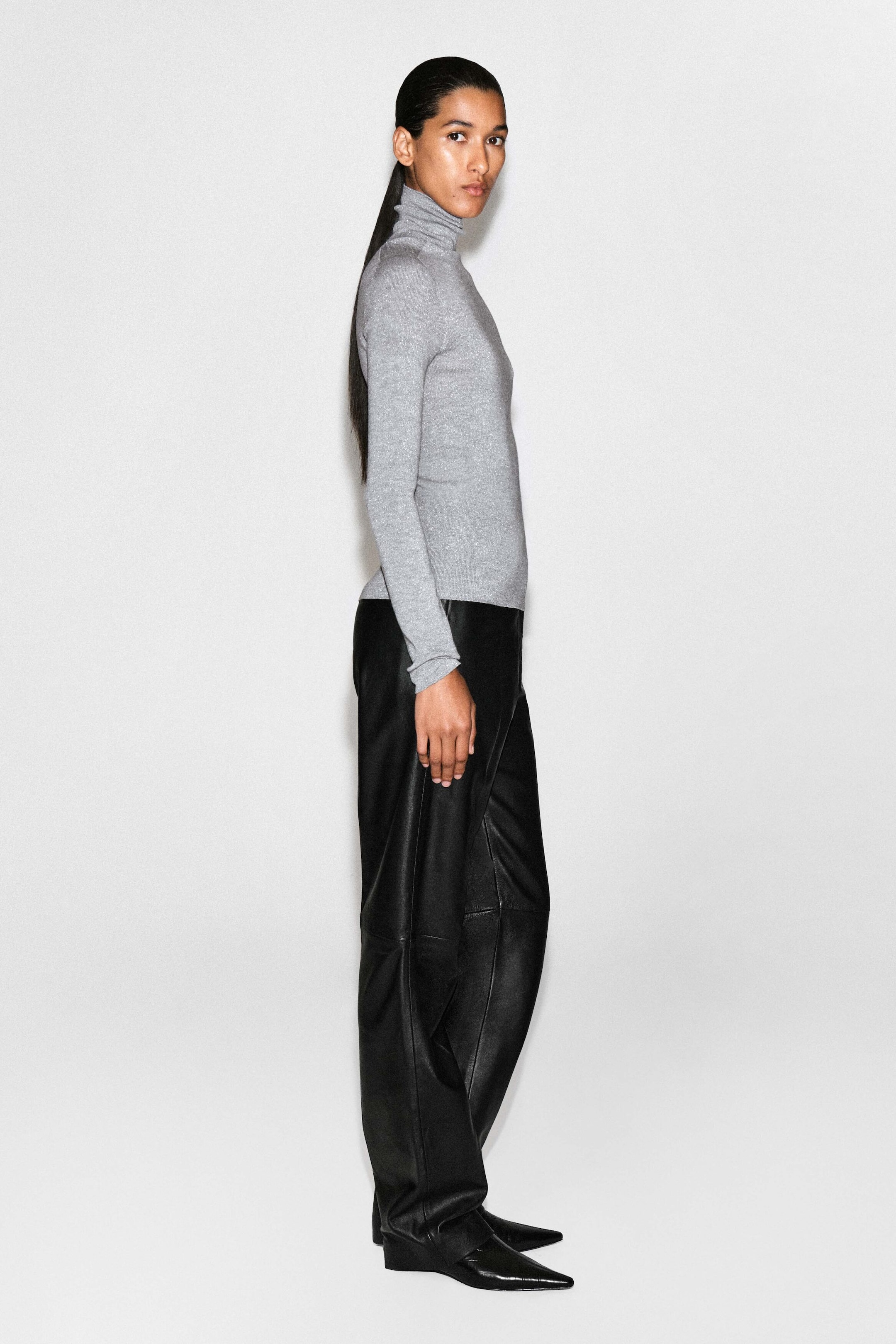 The sparkly seamless wool funnel-neck top is worn in a side-profile view facing right, highlighting its slim fit, long sleeves, and high, structured collar. This light gray fine-knit top features a subtle metallic sheen and is styled untucked over black leather trousers.