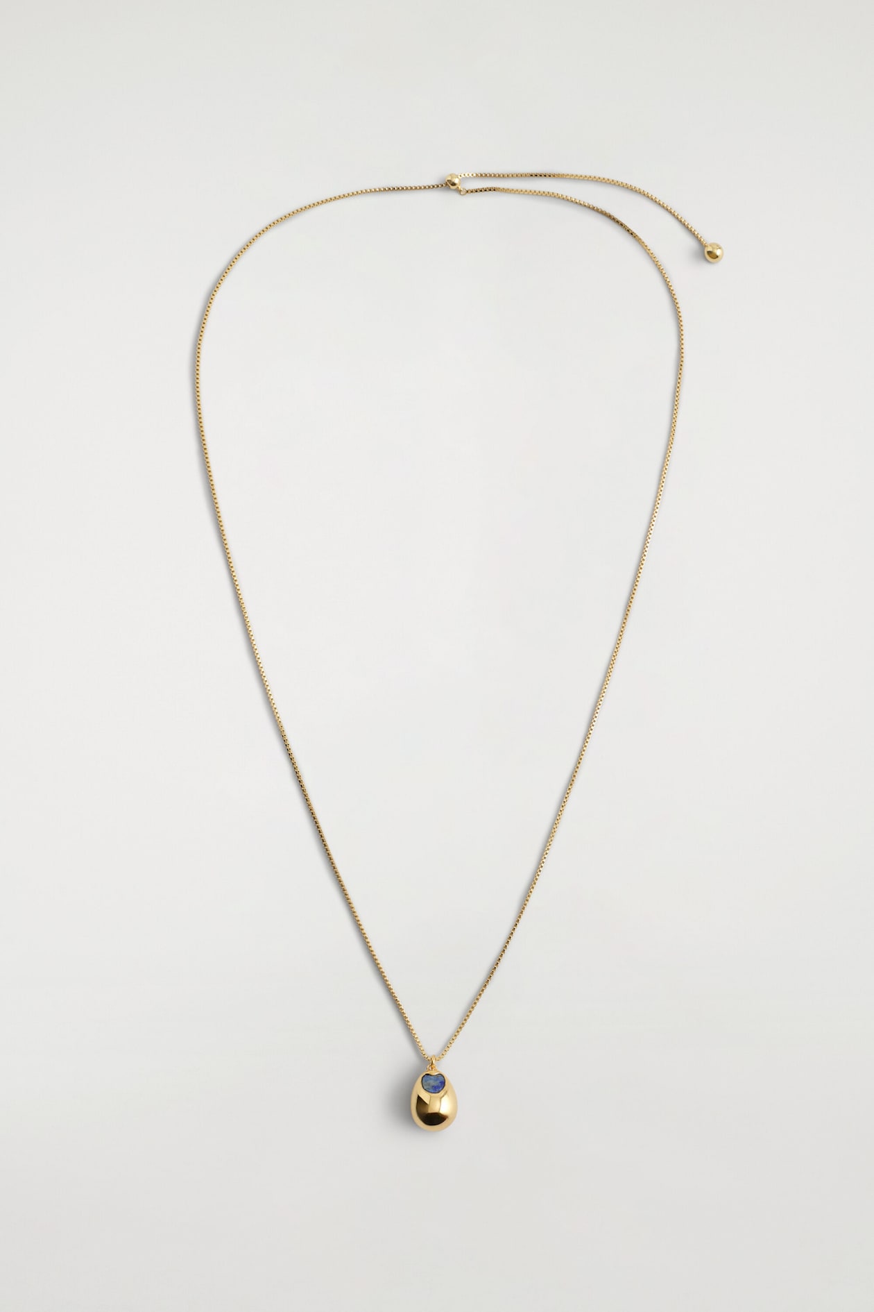 A gold-tone lapis lazuli pendant necklace is laid flat in a front view, featuring a thin box chain holding a smooth, egg-shaped gold pendant. The pendant displays a small inset blue lapis lazuli stone near the top, while the chain ends with an adjustable slider clasp.