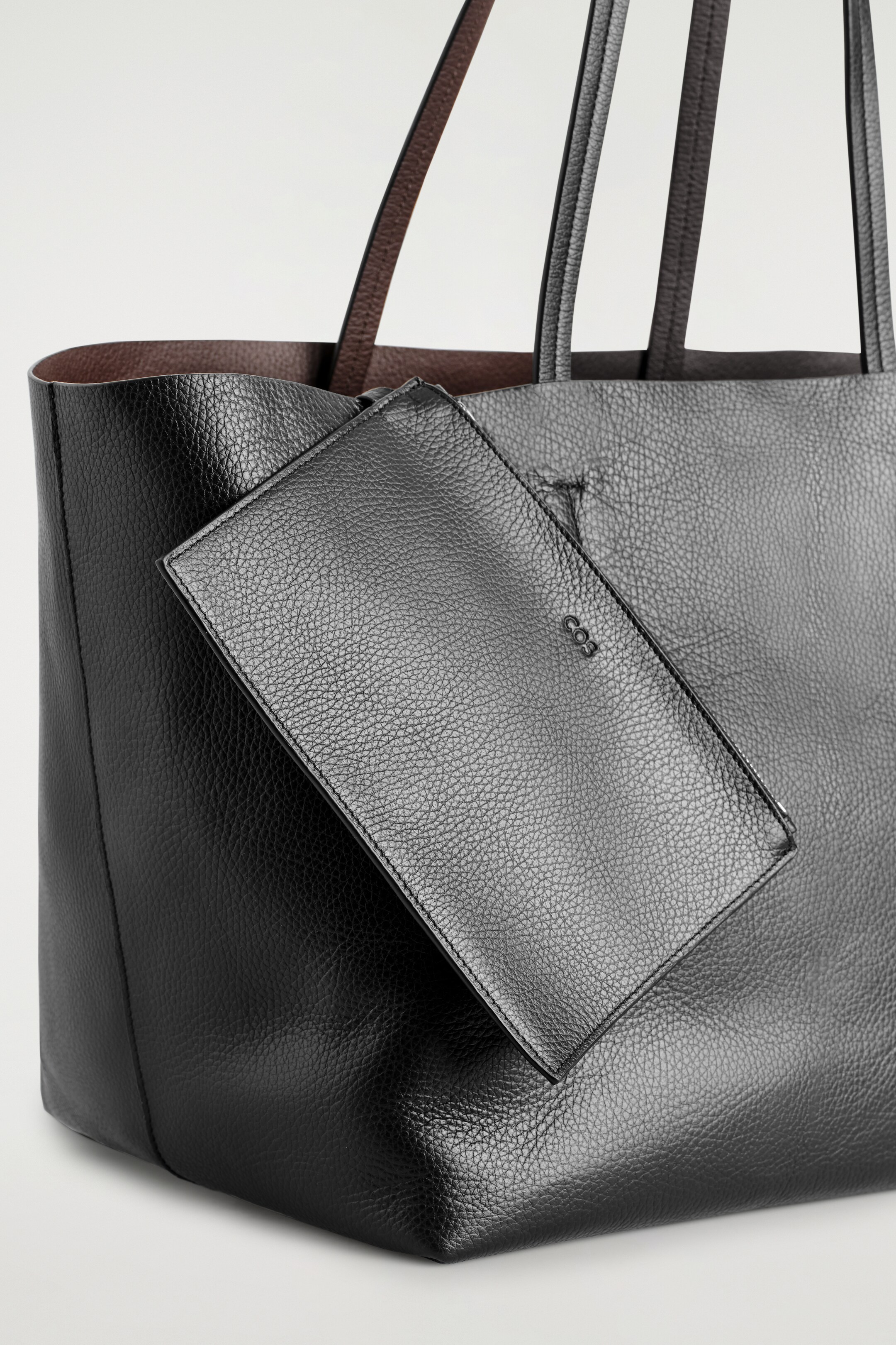 Agrandir l'image: Un sac cabas en cuir noir grainé est présenté sous un léger angle, avec une doublure intérieure marron contrastante et deux anses d'épaule noires. Une petite pochette assortie en cuir noir grainé, estampée avec "COS," est suspendue à l'avant du cabas, montrant son coin inférieur structuré.
