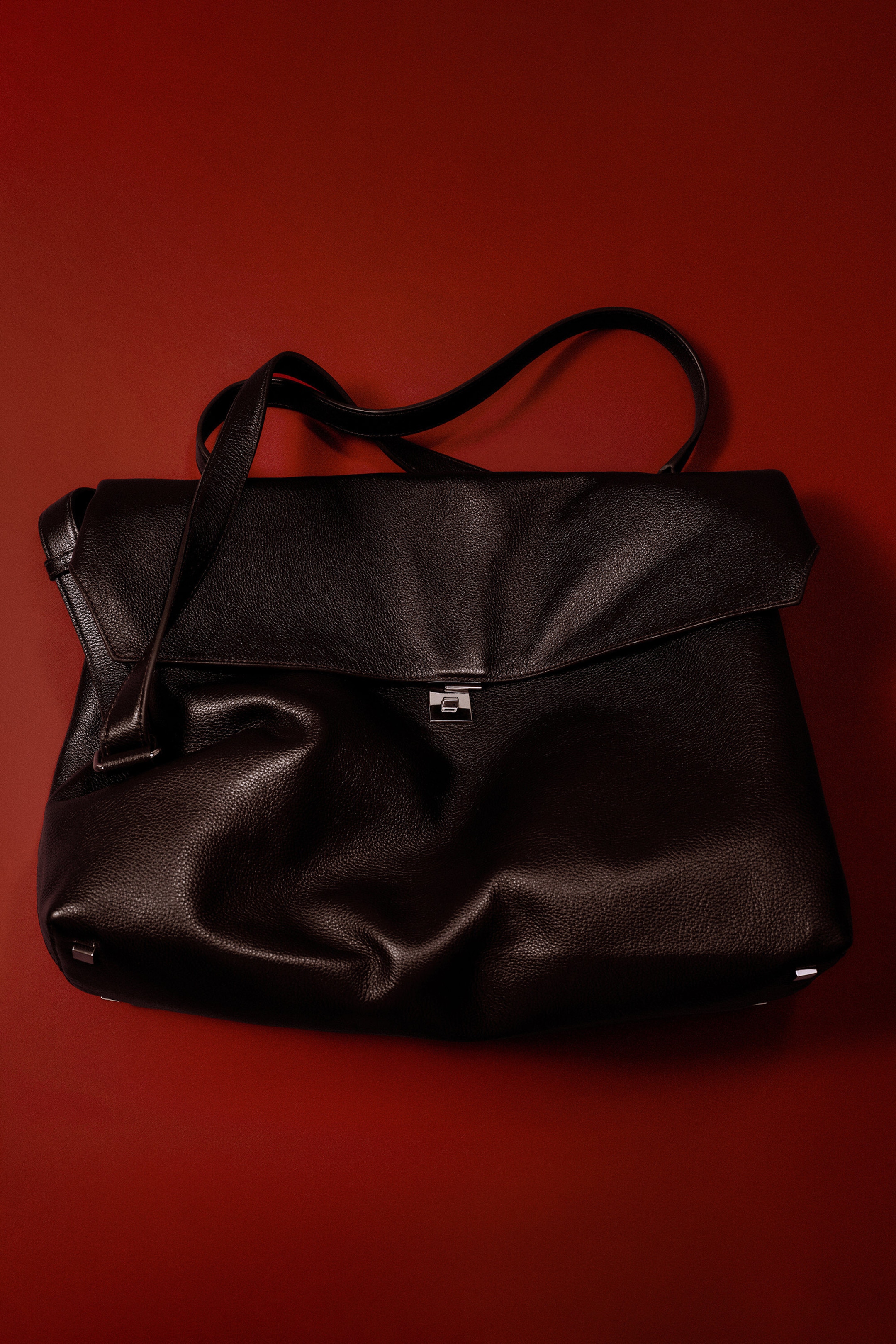 A dark leather handbag is shown front-facing, showcasing a slouchy, folded silhouette with a textured pebbled finish. It features a silver-tone metal lock closure at the centre, a top handle, and a detachable shoulder strap draped over the top.