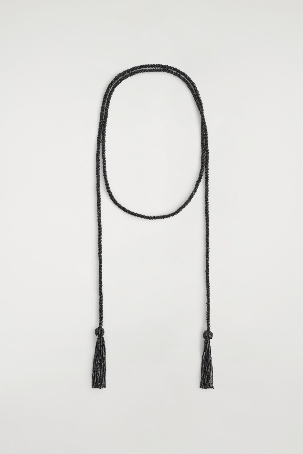 A long black beaded lariat necklace is draped in a loop, with two strands hanging straight down. Each strand features a matching beaded tassel at the end.