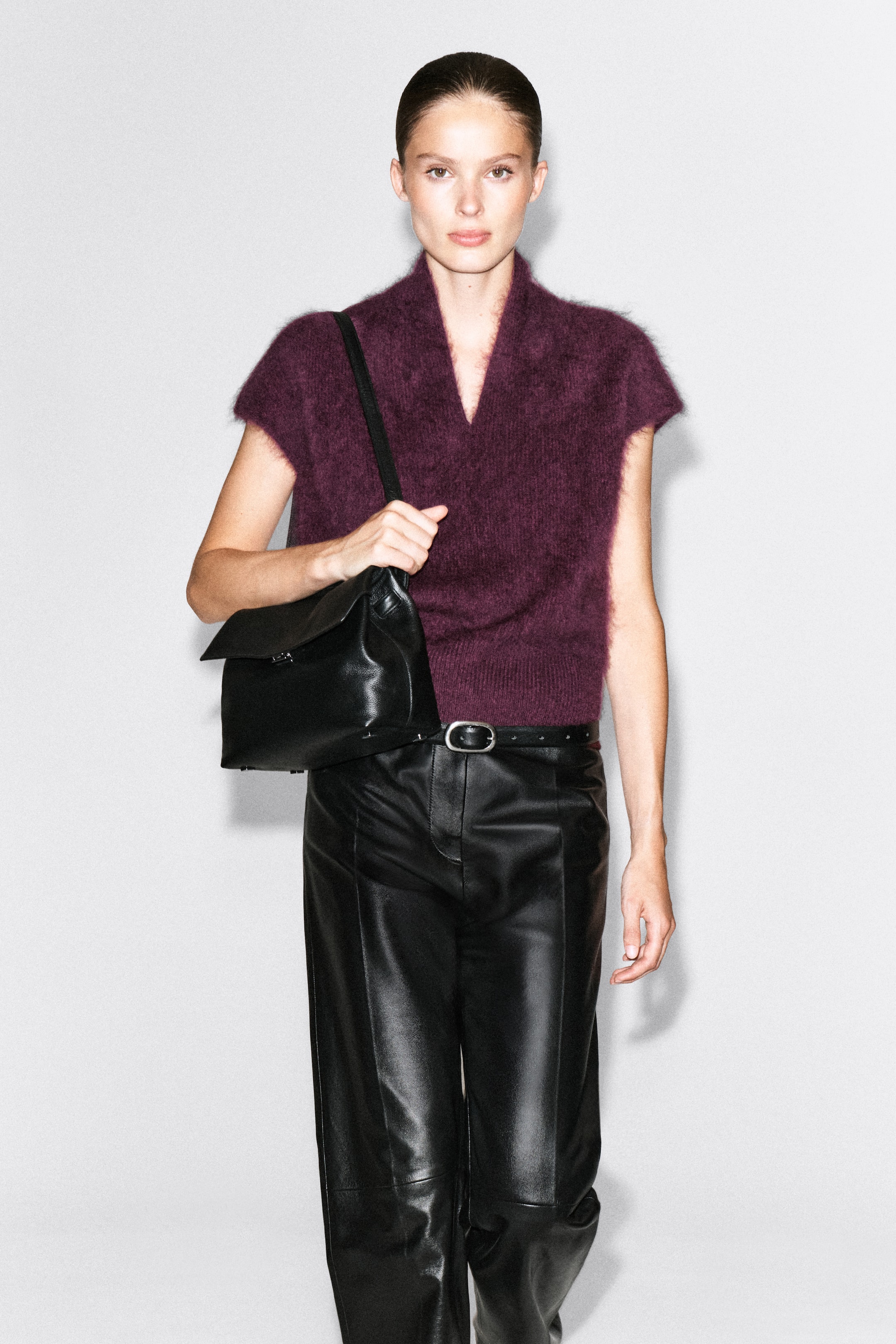 View larger image: A draped brushed merino wool top in a deep plum colour is being worn by the model, featuring a V-neckline, cap sleeves, and a soft, textured finish. The top is styled tucked into black leather trousers, with the model holding the strap of a black leather shoulder bag.