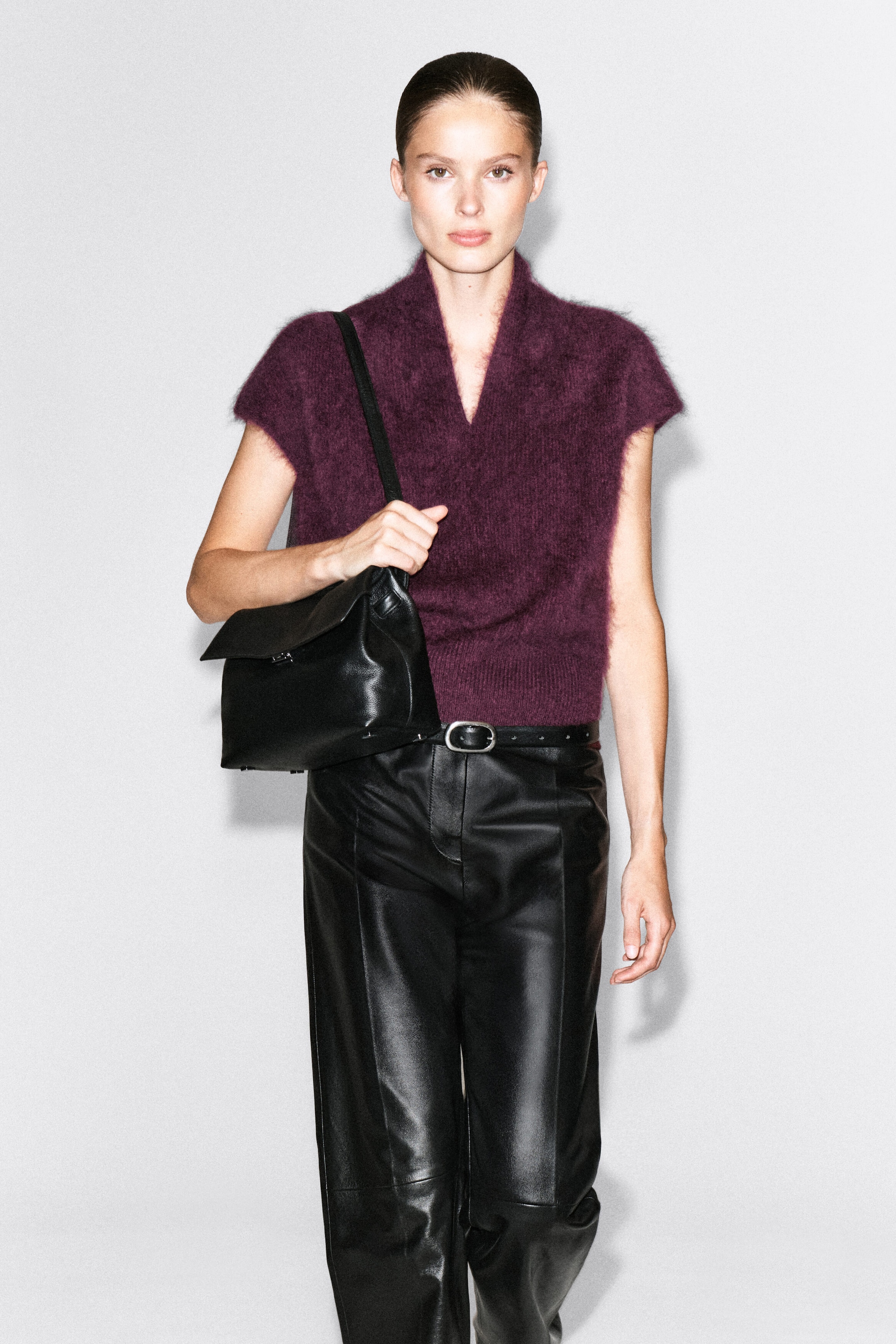 A draped brushed merino wool top in a deep plum color is being worn by the model, featuring a V-neckline, cap sleeves, and a soft, textured finish. The top is styled tucked into black leather pants, with the model holding the strap of a black leather shoulder bag.